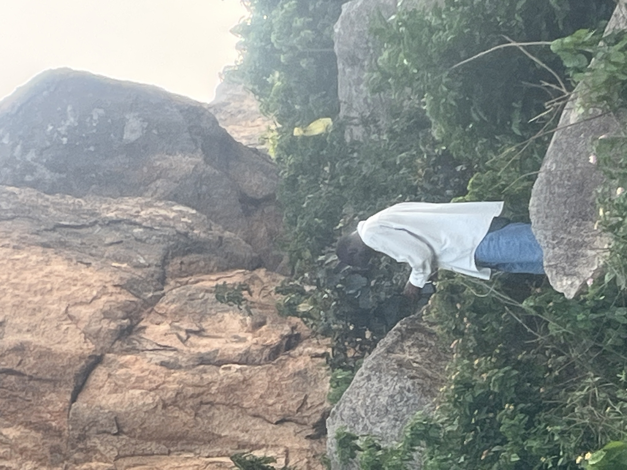 The image captures a rocky, vegetated area within or on the outskirts of Jos, Nigeria. Prominent weathered granite rock formations dominate the background and foreground, interspersed with dense green foliage, including bushes and small trees, some bearing light-colored blooms.
One adult male, of dark complexion, is visible from the back and left side, positioned amidst the rocks and vegetation. He is dressed in a light-colored, long-sleeved shirt and blue trousers. His head is slightly bowed, and his hands are positioned at waist level, engaged in an unclear action.
In the upper right background, a distinct yellow object, appearing to be a plastic bag or piece of fabric, is suspended within the dense greenery. The scene is illuminated by diffused natural light, with a bright, hazy sky visible in the upper right, suggesting either early morning or late afternoon conditions.