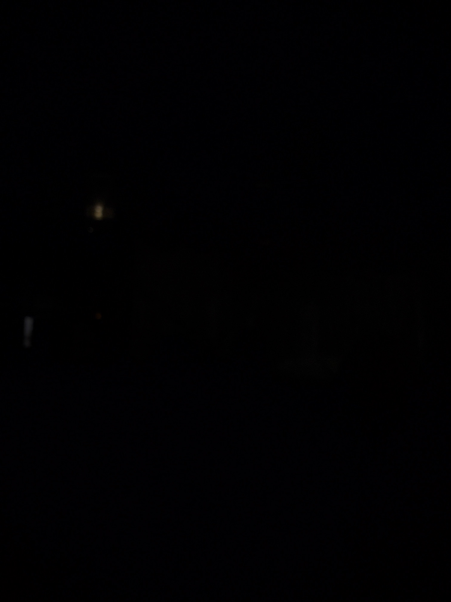 The image depicts a dark, possibly nighttime scene. A small, blurred light source, appearing as a faint yellow glow, is visible in the upper center of the frame. Below this, a blurry, vertical white shape is present on the left side. The rest of the image is predominantly black, suggesting an absence of significant light or detail. There are no identifiable people, objects, or environmental features that would allow for specific identification of the location, including the city of Samara, Russia. The overall impression is one of low light conditions with minimal discernible content.