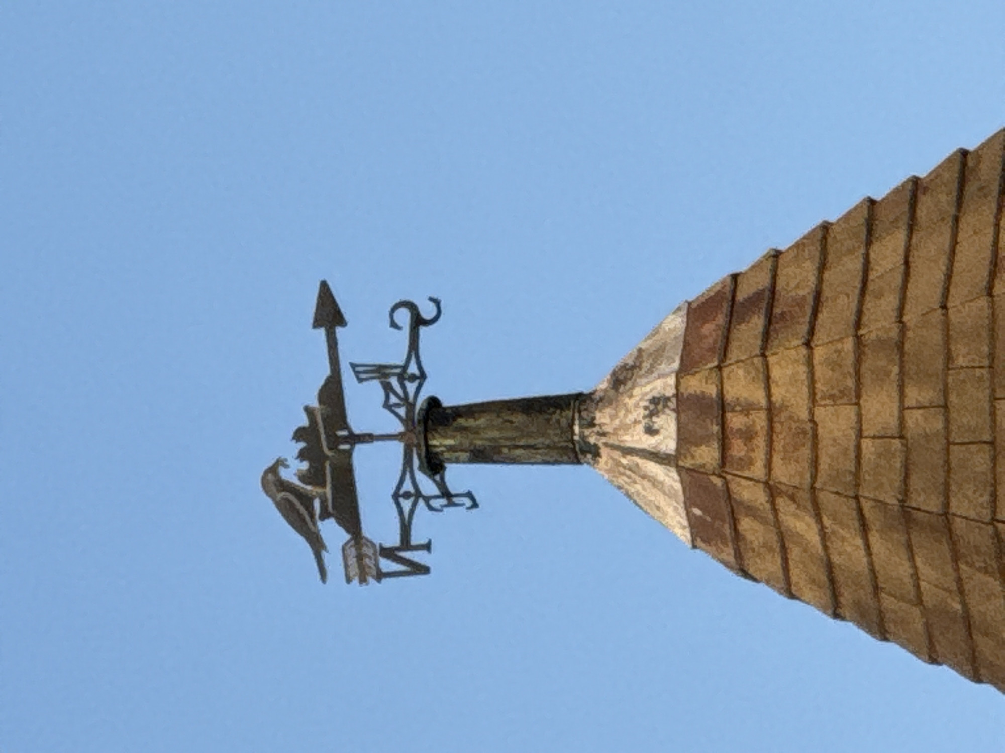A close-up, low-angle shot focuses on the rooftop of a structure. The roof, covered in rectangular, brown tiles, is capped with a cylindrical metal piece. Atop this metal fixture is a weather vane featuring a decorative bird figure and directional arrows (N, S, E, W). The background is a clear, light blue sky. The image does not contain any people, actions, or interactions. The location is unknown.
