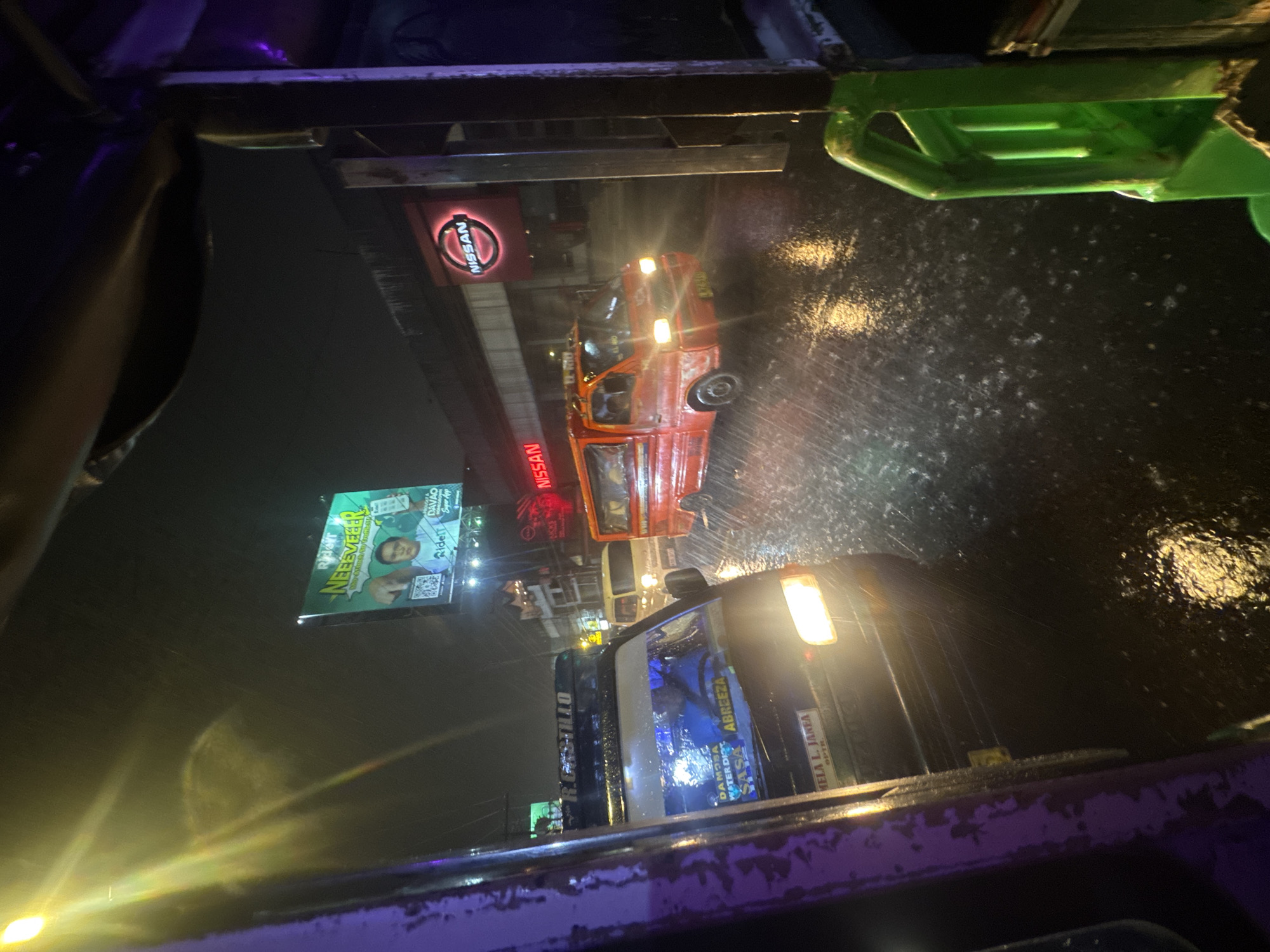 The image shows a street scene in Davao City, Philippines, at night during a heavy rain. The perspective appears to be from inside a vehicle, possibly a tricycle or a similar mode of transport, looking outwards. Several vehicles are visible on the wet, reflective road, including a bright orange van and a dark colored van or SUV. The headlights of the vehicles are illuminated, casting light on the rainy street.
Above the street, there are several billboards and signs. One prominent billboard features a green background with text that reads "NEEEVEER" and an image of a man. Another sign displays the "NISSAN" logo illuminated in red. The overall atmosphere is dark and wet, characteristic of a tropical downpour at night. The presence of multiple vehicles suggests a busy urban environment. The details on the vehicles, such as license plates and labels, are partially obscured by the rain and reflections. One van has text on its windshield that includes "DAMAO WATER DIST" and "ABREEZA". Another sticker on the dark vehicle reads "MELA L. JAREA".