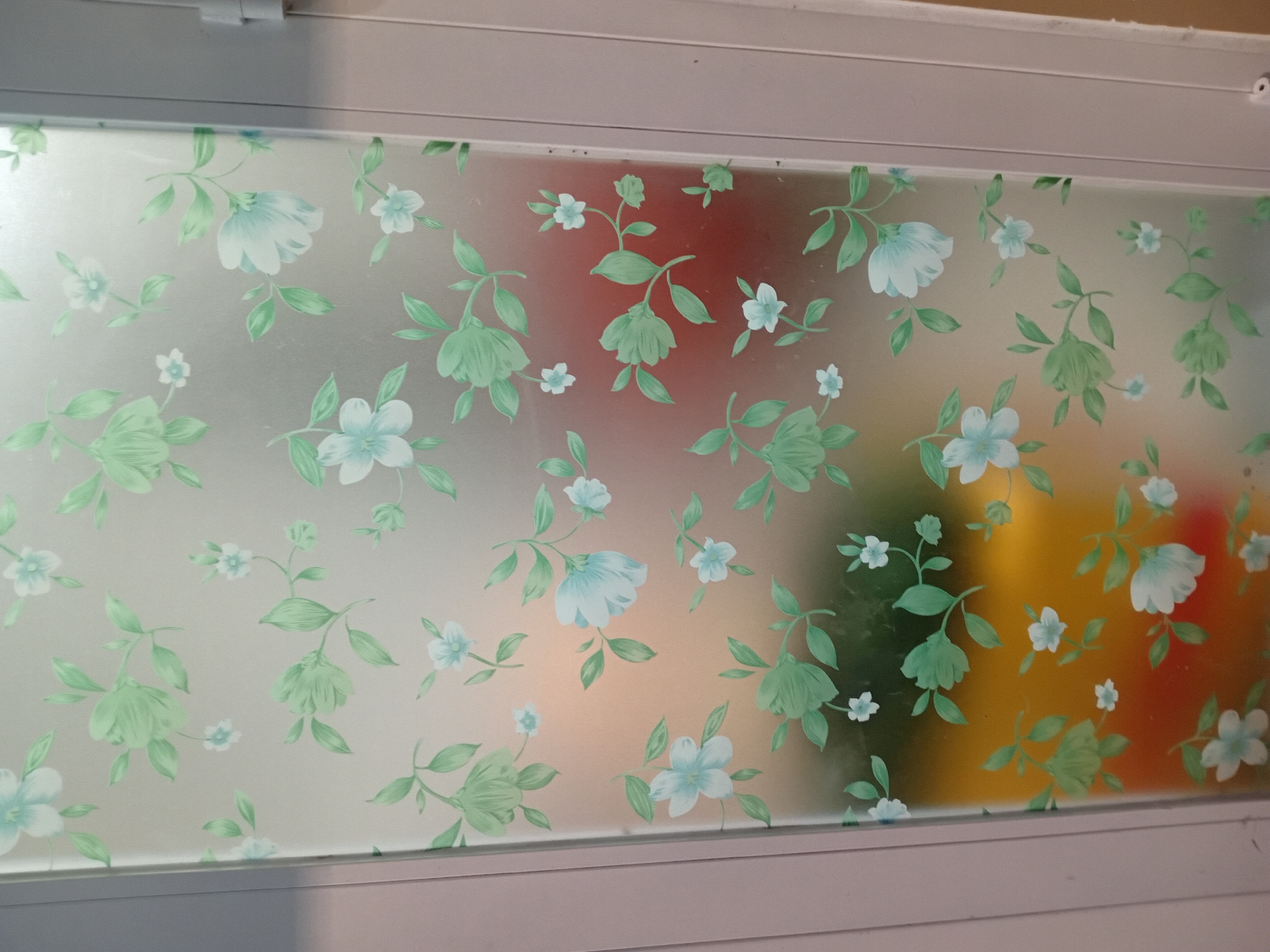 The image displays a frosted glass door with a repeating floral pattern of light blue flowers and green leaves. The glass is translucent, allowing a blurred view of colored objects behind it, which appear to be red, yellow, and green. The door is framed by a light-colored, possibly metallic or painted, door frame. The scene is indoors, but the presence of colored objects behind the glass might suggest it's a partition to a brightly lit area. There is no indication of Đà Nẵng, Vietnam in the image itself.