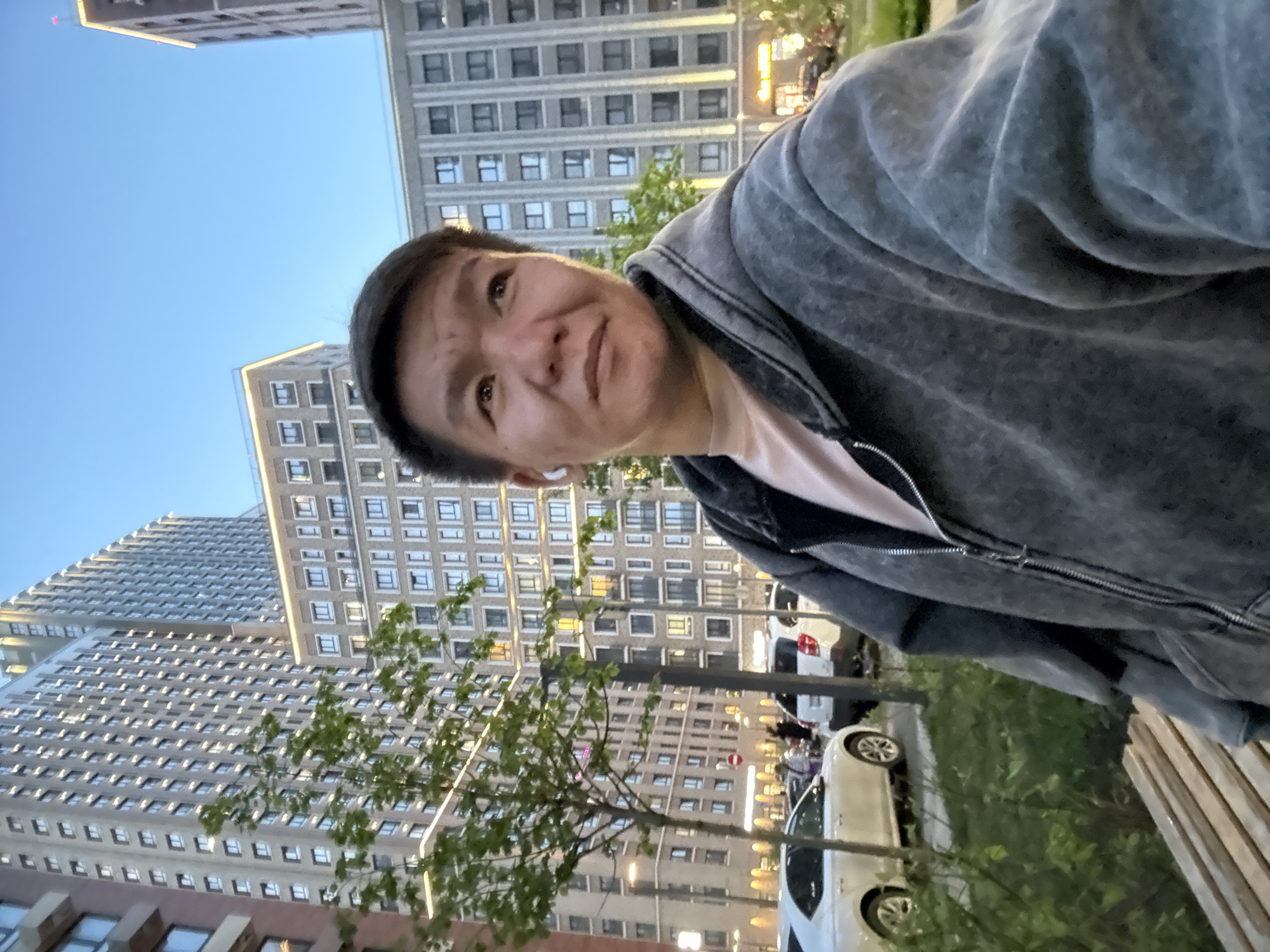 A selfie of a person is taken in an urban environment, specifically near high-rise buildings in Moscow, Russia. The person is wearing a grey hooded jacket over a pink shirt. Buildings with numerous windows are in the background. There are trees and parked vehicles visible. The time of day appears to be dusk or early evening.
