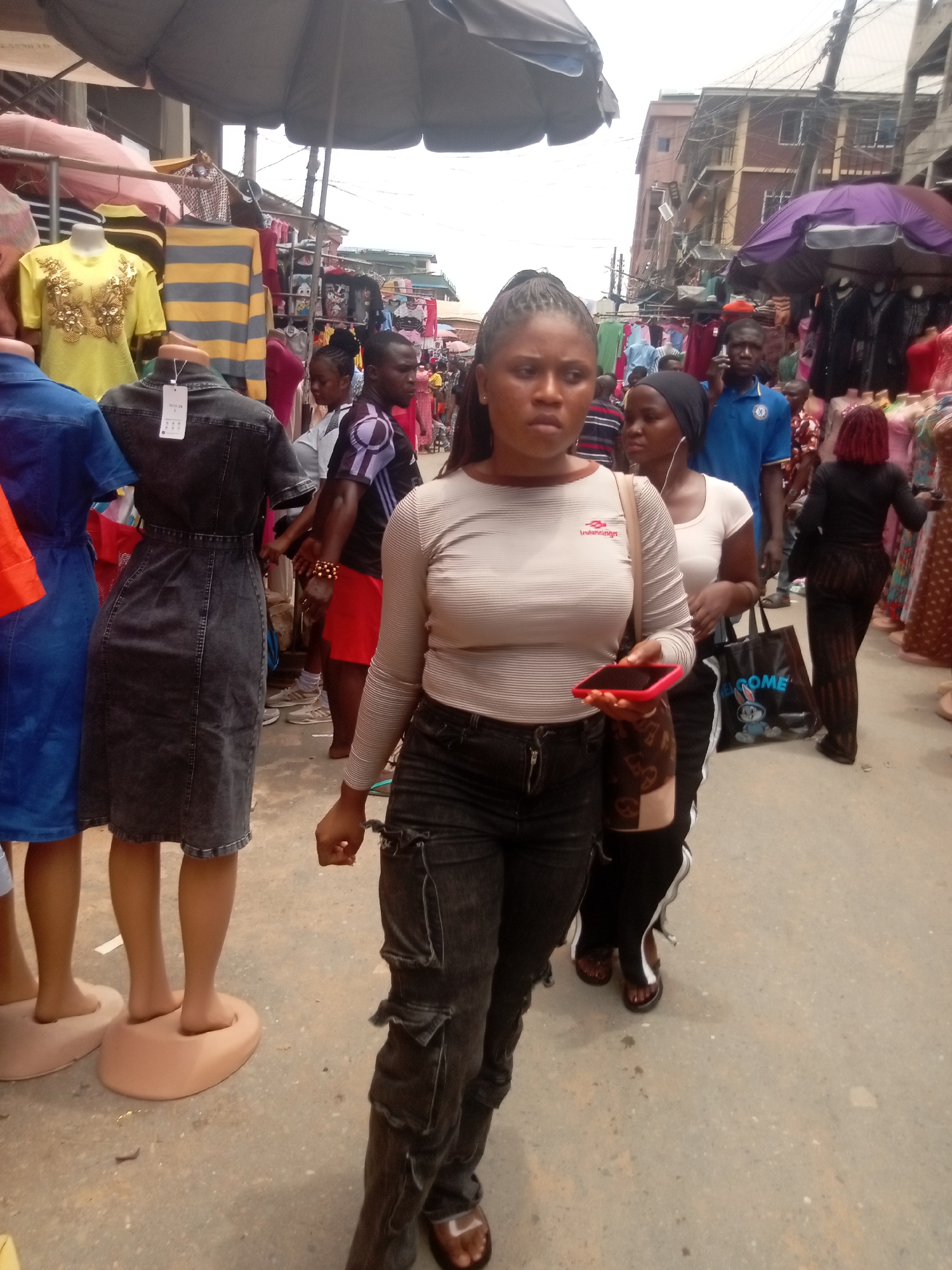 The image captures a bustling scene in Fegge, Nigeria, likely a market street. The foreground is dominated by a woman with dark hair in a ponytail, wearing a striped long-sleeved shirt and dark, possibly denim, pants. She holds a cell phone with a red case. Another woman in a white shirt with black pants is partially visible behind her. Both are walking through the crowded space.
Clothing is a prominent feature, with mannequins displaying various garments, including a blue dress, a grey denim dress, and a yellow top. Other people are milling around, with clothing displayed on racks or stands.
The environment appears to be a crowded outdoor market with umbrellas providing shade over the vendors. The ground is concrete and shows signs of wear. Buildings are visible in the background, suggesting a dense urban area. Electrical wires are strung overhead.