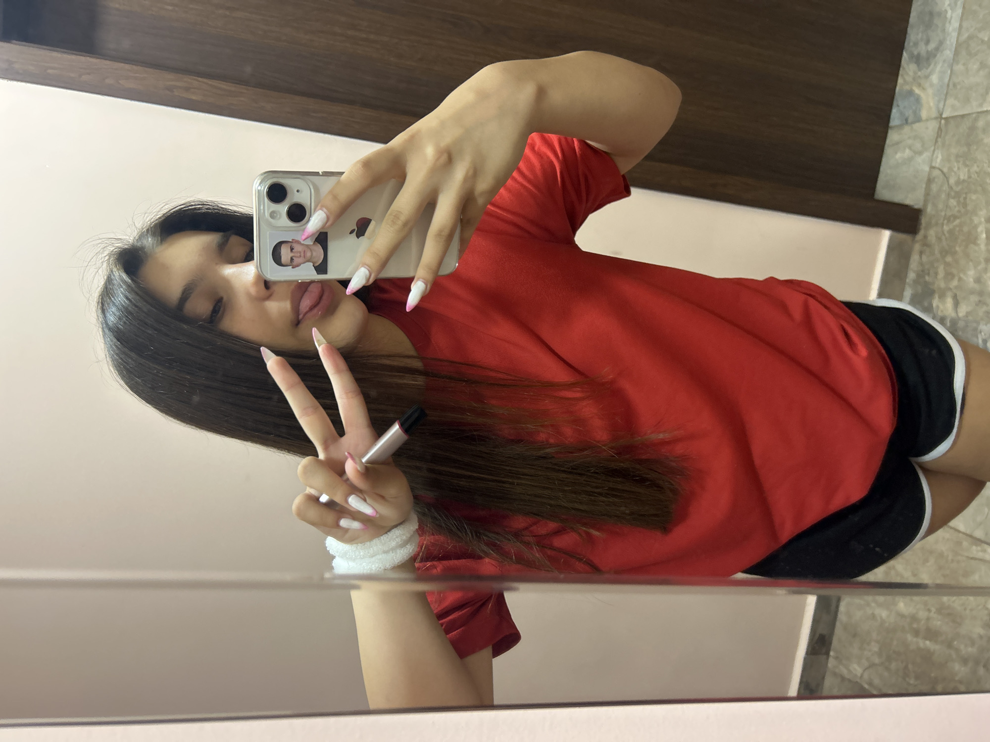 A young woman is taking a selfie in a mirror. She is wearing a red t-shirt and black shorts. Her nails are painted white with pink tips. She is sticking her tongue out and holding up two fingers in a peace sign. A small photograph of a man is attached to her phone case. The background appears to be a room with tiled flooring and a wooden door. This image was taken in Shumen, Bulgaria.