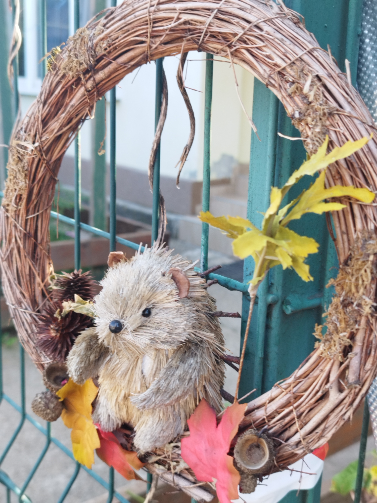 An autumnal wreath made of twigs is hanging on a teal metal fence in Bucharest, Romania. A plush hedgehog figure is attached to the wreath, nestled among pine cones, acorns, and colorful fall leaves. The background shows a residential building and hints of greenery.