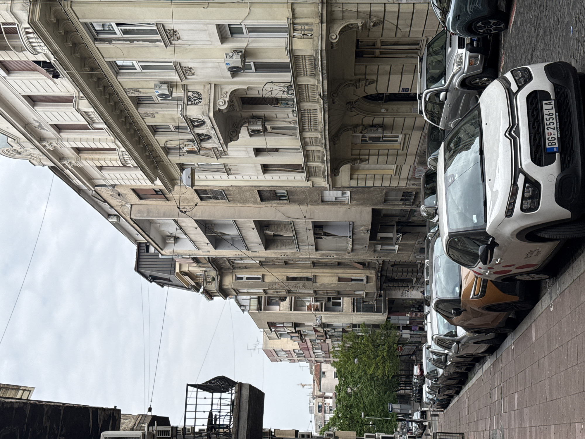 The image depicts a street scene in Belgrade, Serbia. The scene showcases a row of parked cars along a brick sidewalk. The vehicles range in color from white to gray and brown. The foreground features a white Citroen with a Serbian license plate "BG 2556-LA." Adjacent to the sidewalk, tall buildings dominate the view. These buildings display a variety of architectural styles. There are numerous windows, some fitted with air conditioning units. The sky above is overcast and gray. In the middle ground, a tree is partially visible among the buildings. In the foreground there are visible elements such as a metal construction of some kind and a possible flat roof, suggesting an older or industrial area in the city.