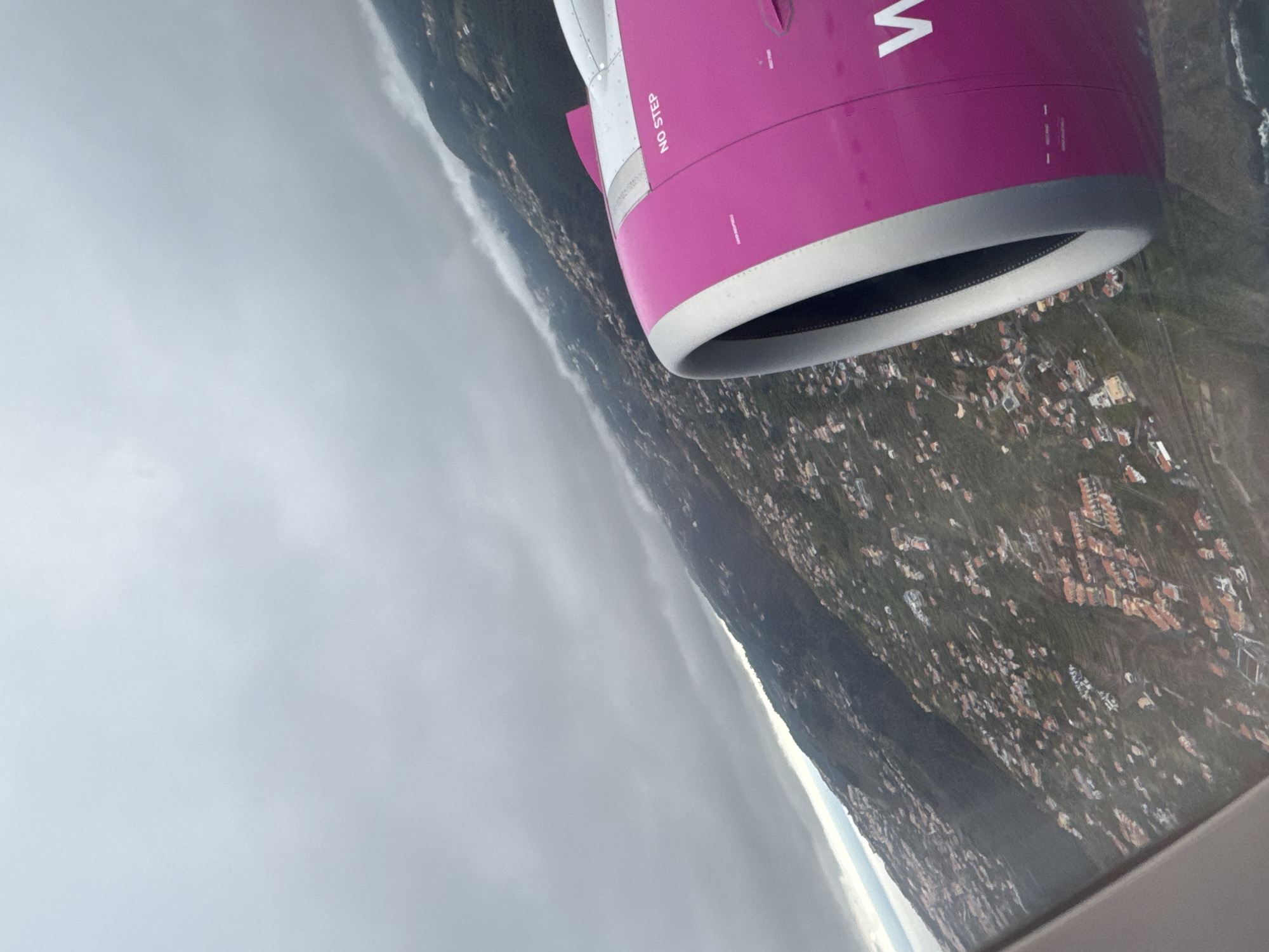 The image, taken from inside an airplane, shows a view out the window, including the wing and engine. The engine is magenta with a silver trim and displays the logo "W". The ground below shows a cityscape of Sosnowiec, Poland, with buildings interspersed among green areas. A cloud bank is visible in the distance.