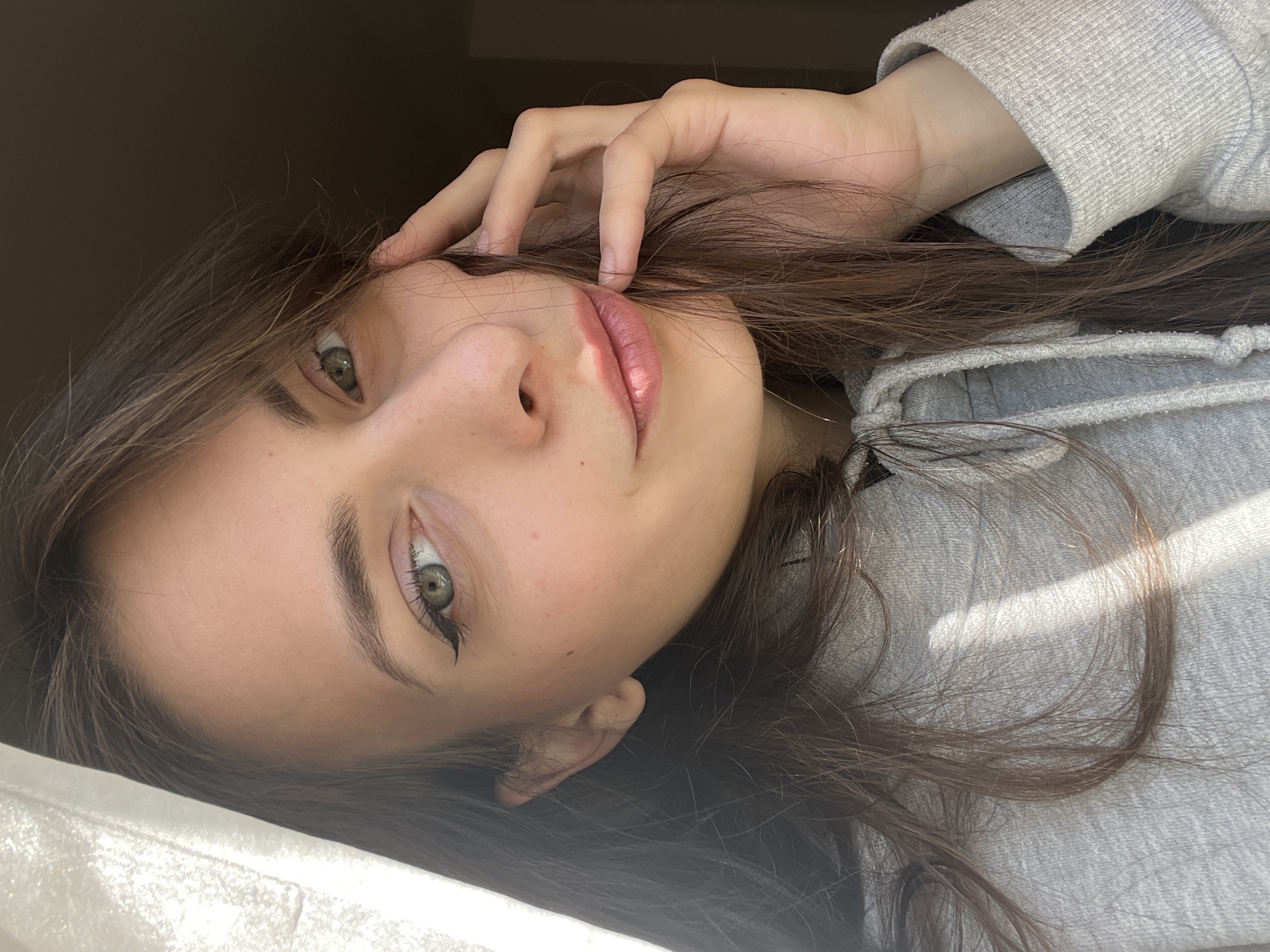 A young woman with light skin, green eyes, and brown hair is looking directly at the camera. Her head is tilted to the right. She is wearing a grey hooded sweatshirt. Her right hand is near her face, with her fingers slightly touching her forehead. The background is blurry but appears to be an indoor setting with natural light coming from a window. There is not sufficient information available to determine the location in the city of Krasnoyarsk, Russia.