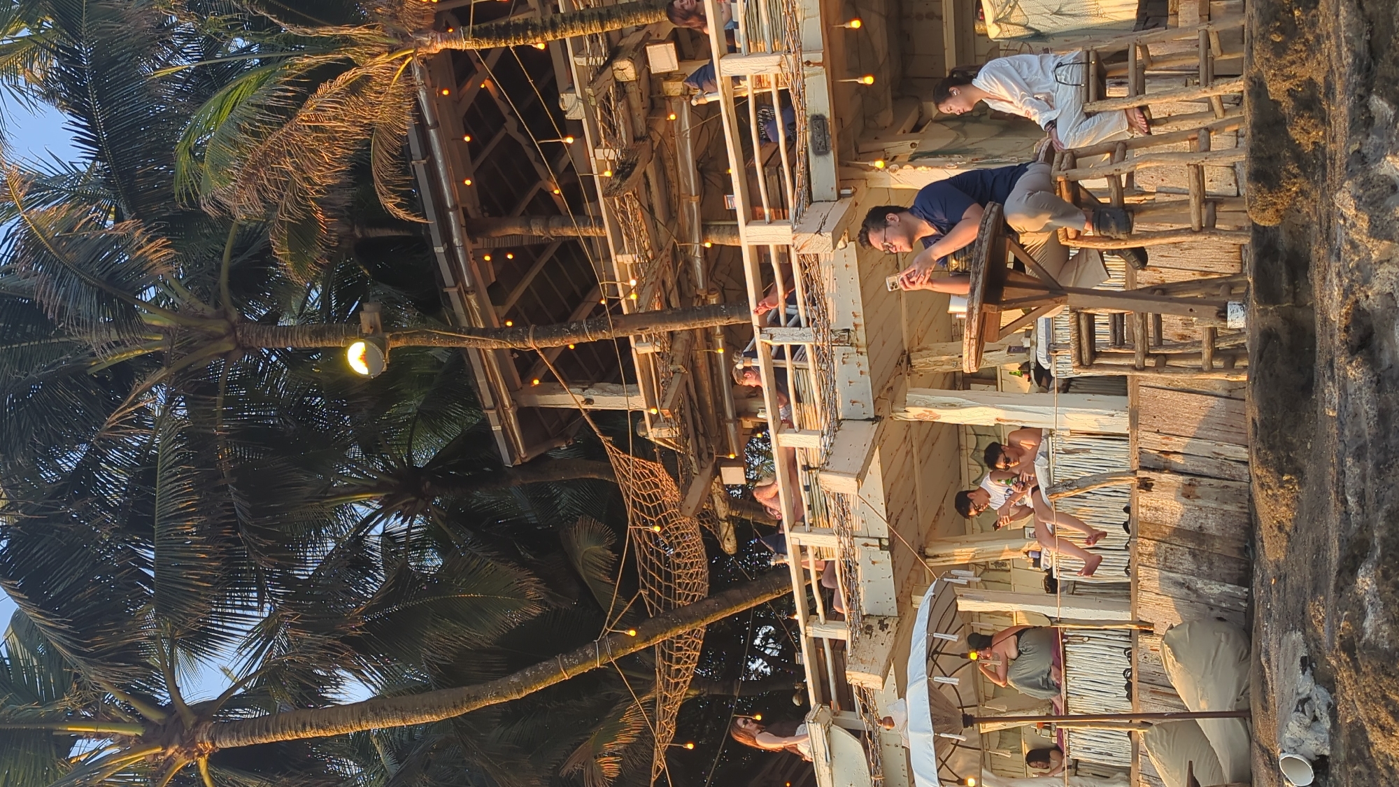 The image shows a multi-level wooden structure, possibly a restaurant or lounge, in North Kuta, Indonesia. Several people are present on different levels of the structure. One person is looking at their phone. Palm trees with string lights surround the structure. The time of day appears to be evening. The structure has wooden railings and supports. Some people are seated at tables, while others are standing.