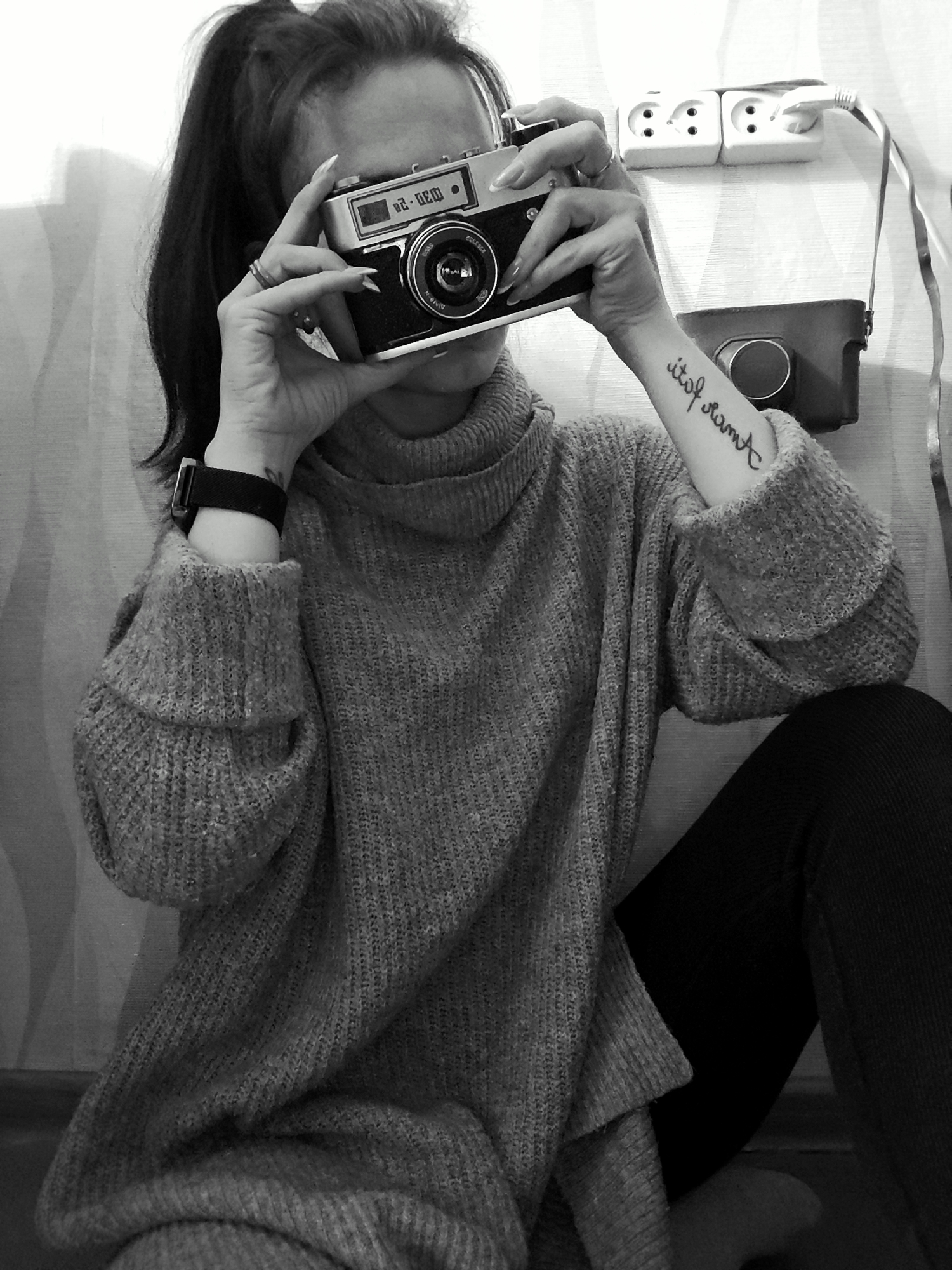 This monochrome image captures a person, likely female, holding a vintage film camera in an indoor setting in Фоки, Russia. The subject is seated, wearing a light gray, chunky knit turtleneck sweater and dark trousers. Their dark hair is styled in a ponytail.
The person holds a classic rangefinder camera with both hands, positioned in front of their face, obscuring most of their features as if looking through the viewfinder or preparing to take a shot. The camera has a metallic top plate and black body, with visible Cyrillic text, possibly "ШО0-20," on its exposure counter or film type dial. The subject's hands show long, manicured fingernails, and a ring is visible on one finger. A dark-strapped watch or bracelet is worn on the left wrist.
A notable detail is a tattoo on the subject's right forearm (from the viewer's perspective), which appears reversed, displaying the Cyrillic phrase "этот память," translating to "this memory."
The background features a light-colored wall with a subtle vertical textured pattern. Mounted on the wall are two white double electrical outlets, one of which has a white power plug inserted, with its cable extending downwards. Below the outlets, a dark leather camera case or bag is visible, resting against the wall. The overall scene suggests a quiet, focused moment related to photography.