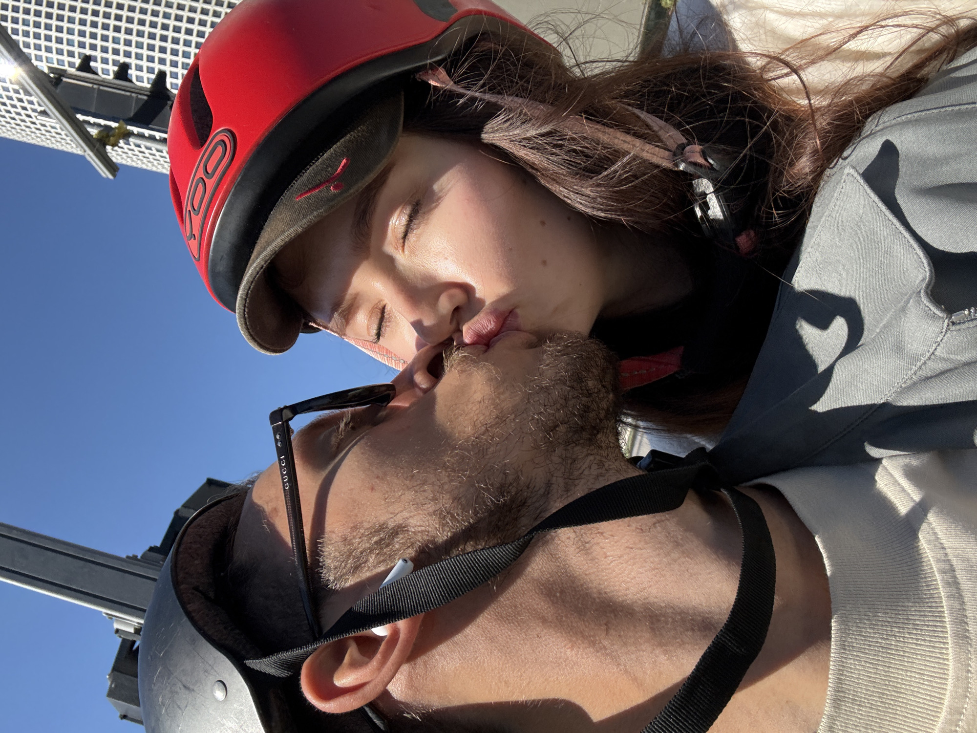 A photograph, taken from a low angle, shows two individuals kissing. The person on the left is wearing a black helmet with a chinstrap, along with black-rimmed glasses with the word "GUCCI" visible on the temple. A white earbud is visible in their ear. The person on the right is wearing a red helmet. The background includes a clear blue sky and a metallic structure with a gridded pattern. The photograph does not contain any information to confirm it was taken in Tel-Aviv, Israel.