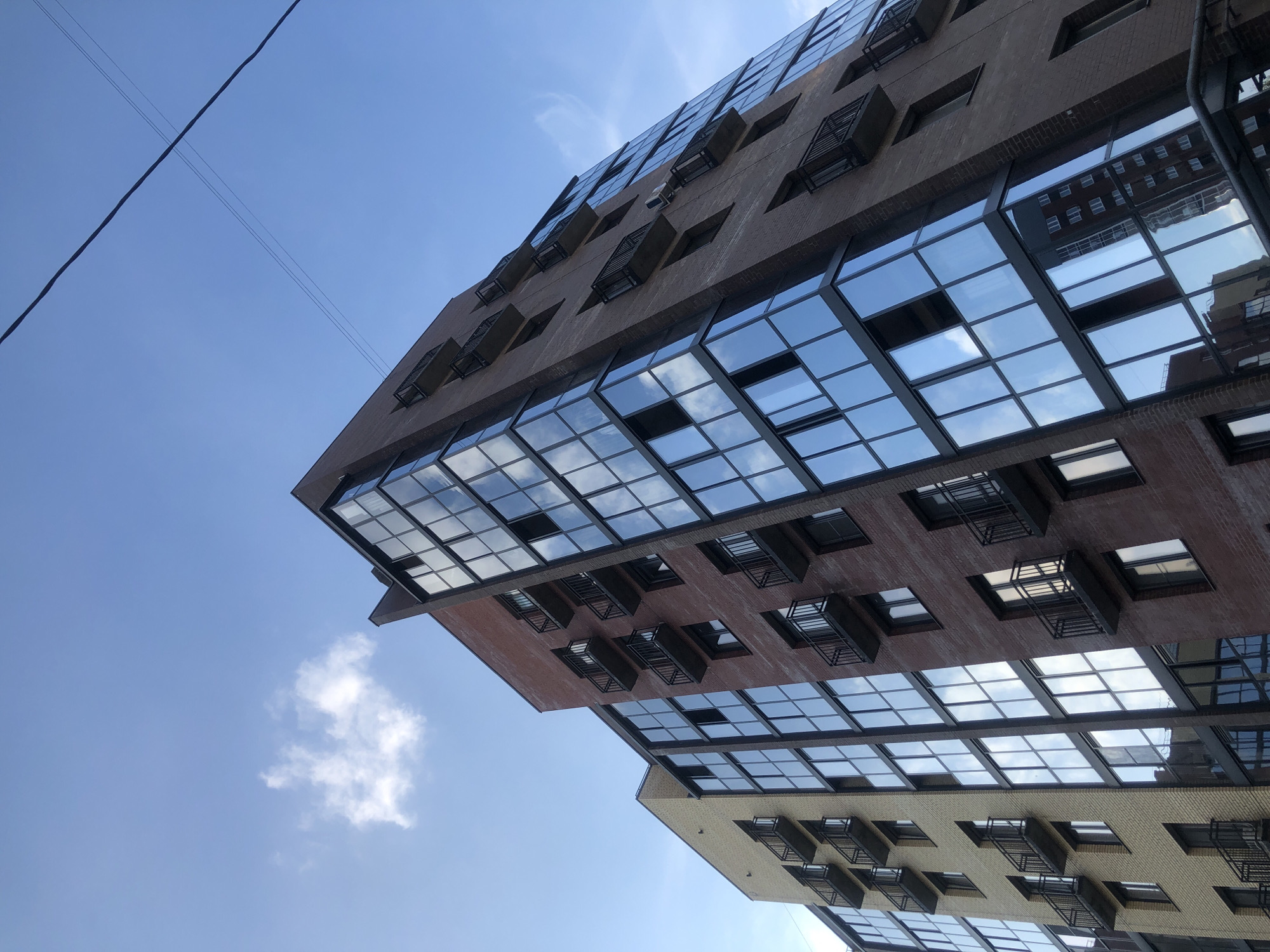 The photograph shows a modern, multi-story apartment building with a facade of brown brick and extensive glass windows, some of which are balconies. The building is seen from a low angle, looking upwards towards a blue sky with a few wispy clouds. A power line stretches across the top of the frame. The architectural style suggests a contemporary urban setting.