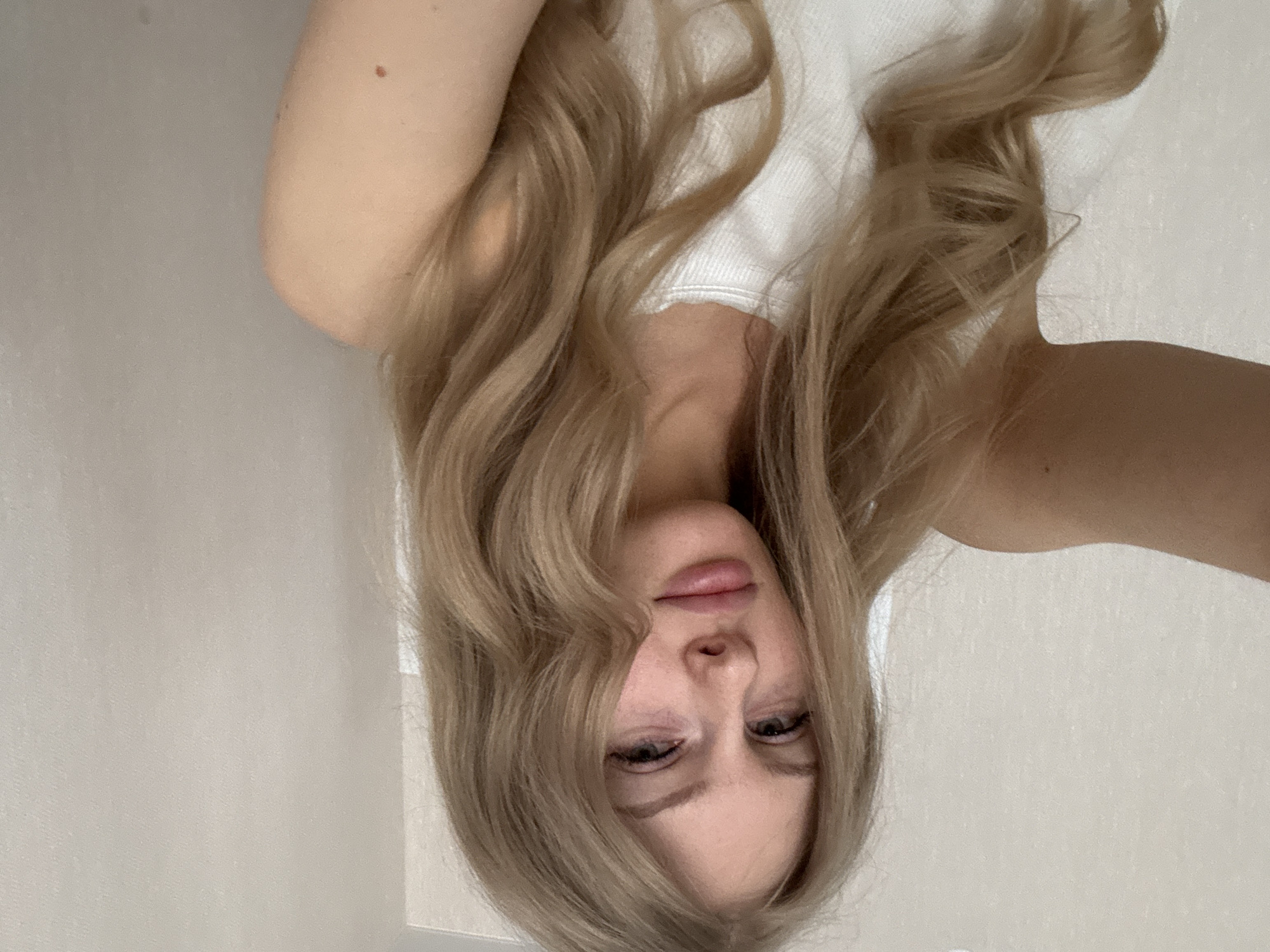A pale-skinned woman with light blonde wavy hair is seen from a slightly overhead perspective. She is wearing a white top. The background appears to be a light beige wall. There is no evidence to suggest the location is Perm, Russia.