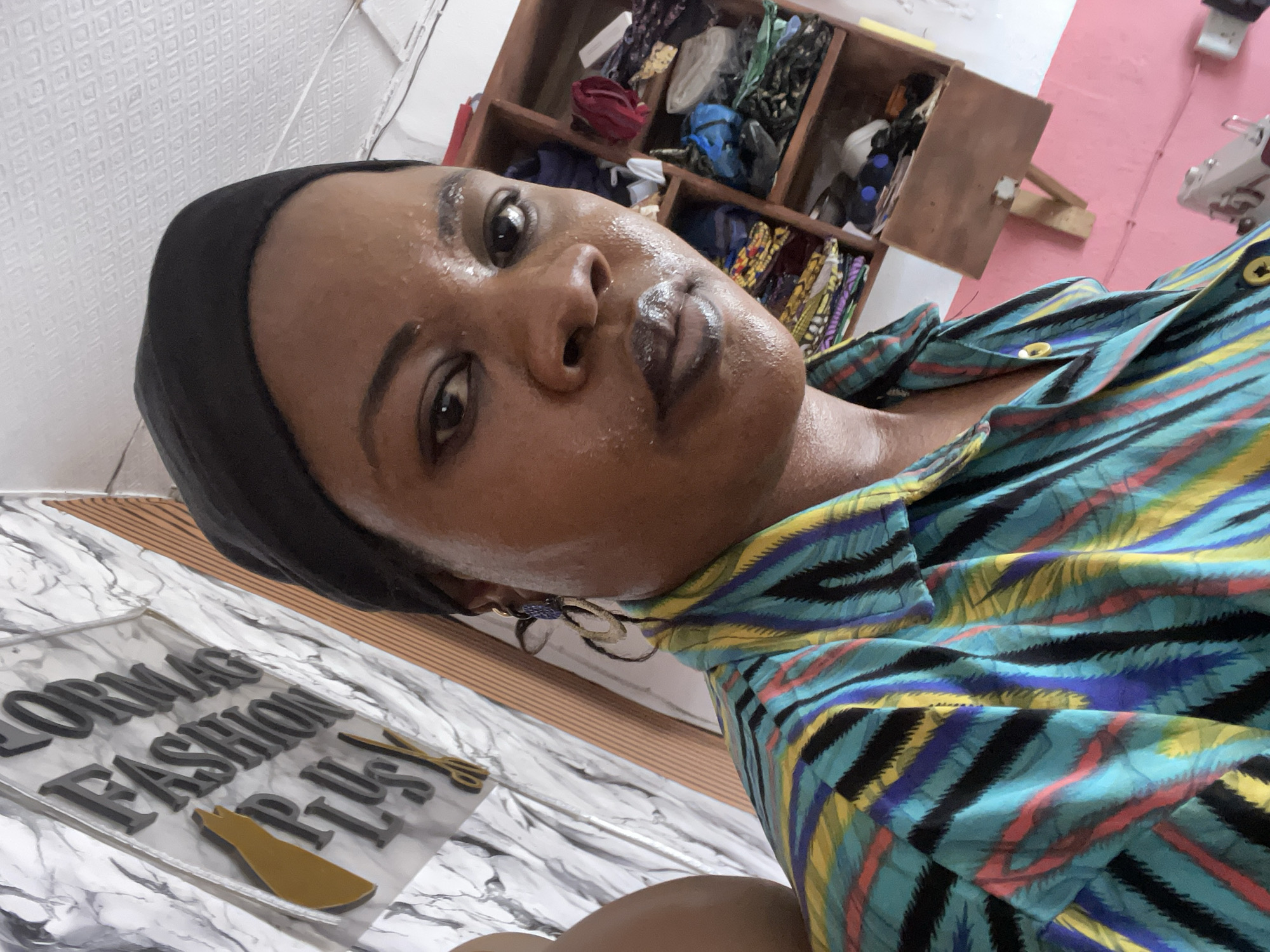 A close-up, low-angle portrait shows a Black woman with damp skin and a black head covering. She wears a buttoned shirt with a striped pattern of black, blue, yellow, and red on a turquoise background. Behind her, a partially visible wooden shelf unit displays textiles. To her left, a white surface with a marble-like pattern features lettering that reads "LORMAC FASHION PLUS". The background includes a white wall with a patterned surface, a pink wall section, and a partial view of a sewing machine. The specific location in Badagry, Nigeria, is not discernible from the image alone.