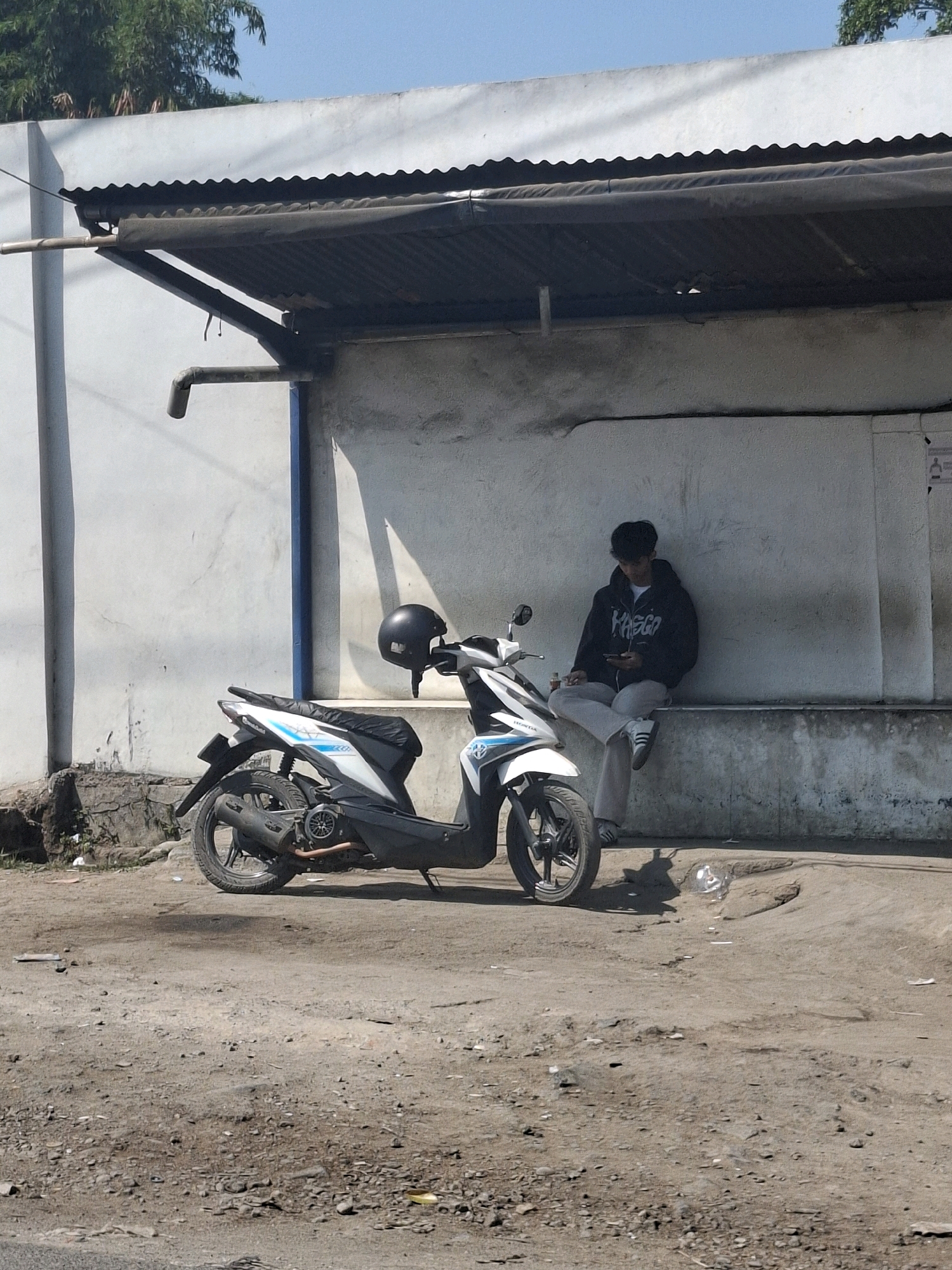 A young man in a black hoodie and light-colored pants sits on a low concrete ledge against a rough white wall. He is looking down at his phone. To his left is a white and blue scooter, its black helmet resting on the handlebars. The scooter is parked on a dirt and gravel surface, with scattered debris. A corrugated metal overhang with a gutter runs across the top of the image. The scene appears to be in an outdoor area, possibly a roadside shelter or an informal commercial space, in Kutasirna, Indonesia, indicated by the tropical vegetation visible in the upper left corner and the overall arid environment.