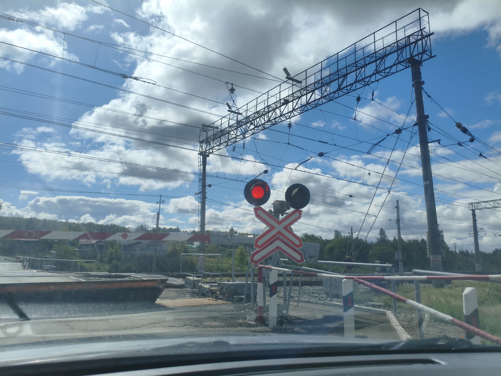 The image depicts a railway crossing in Smolino, Russia, on a cloudy day. A red railway signal light is illuminated, indicating an approaching train or a closed crossing. A red and white striped barrier is lowered across the road. Overhead power lines for the electric railway stretch across the sky, supported by metal structures and concrete poles. In the background, trees and buildings are visible. The image appears to be taken from inside a vehicle, looking out through the windshield.