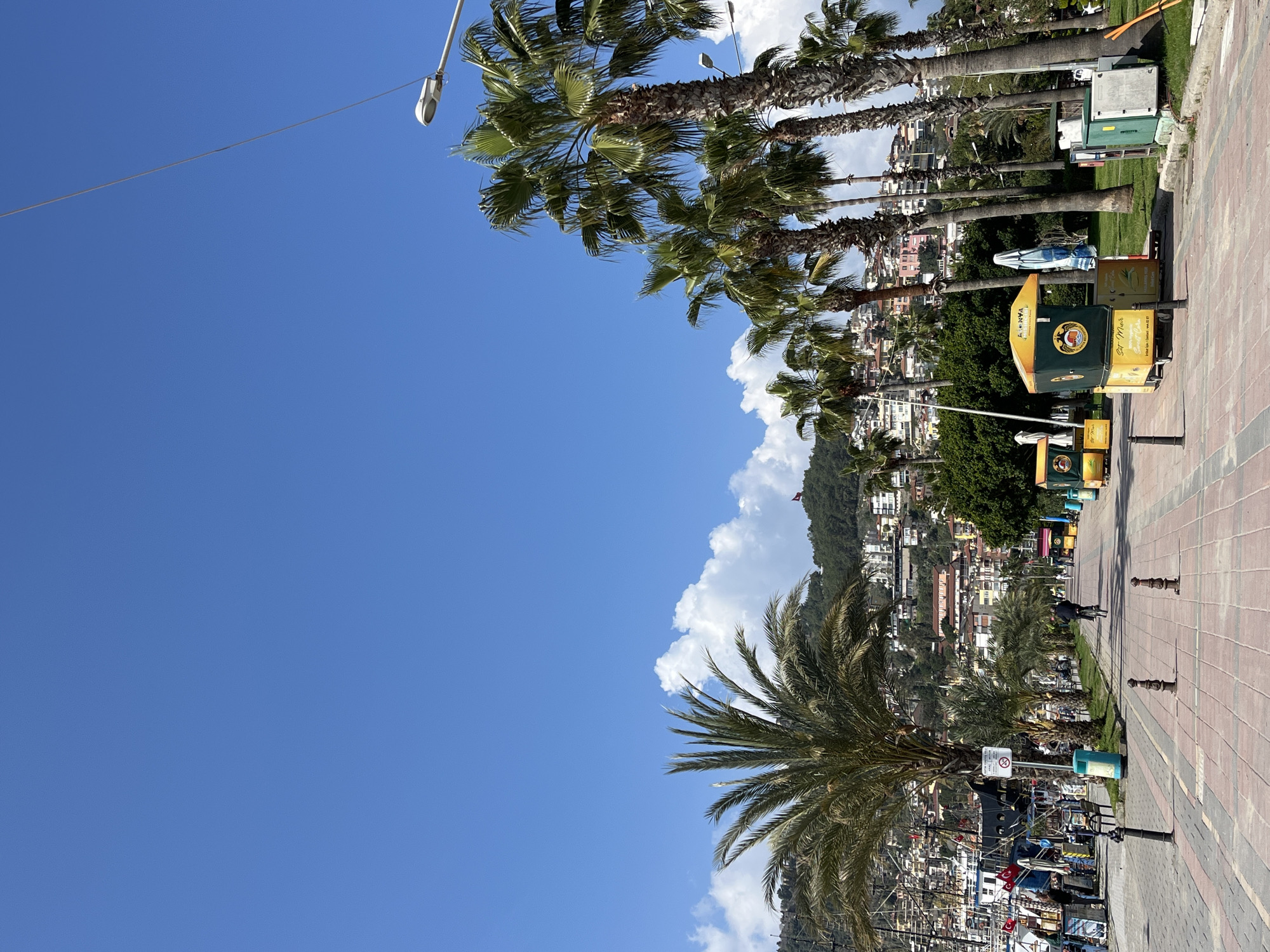 The image depicts a waterfront promenade in Alanya, Turkey, on a clear, sunny day. The scene is dominated by a wide, paved walkway constructed from red and tan interlocking pavers, extending from the foreground towards the background. Dark bollards are spaced along the left edge of the promenade.
To the immediate left, a harbor is visible, populated by numerous docked boats, some adorned with Turkish flags, and flanked by temporary stalls or vendor setups with several individuals standing nearby. A line of palm trees runs parallel to the promenade, separating it from the harbor.
To the right of the promenade, a grassy area borders the path, featuring additional rows of tall palm trees. Several mobile vendor kiosks, yellow and green in color with "ALANYA" branding, are positioned along this side, accompanied by folded blue and white striped umbrellas. Electrical utility boxes are visible near the kiosks further down the path. Streetlights are mounted on poles among the palm trees.
One individual is observed riding a bicycle away from the viewer on the promenade, approximately midway down the path. Another pedestrian is visible further in the distance.
The background is dominated by a steep, green hillside densely covered with multi-story residential buildings in varying pastel and earth tones. On a prominent peak of this hill, a large Turkish flag is visible. The sky is bright blue with scattered white cumulus clouds concentrated above the hills. Shadows are distinct, indicating strong sunlight.