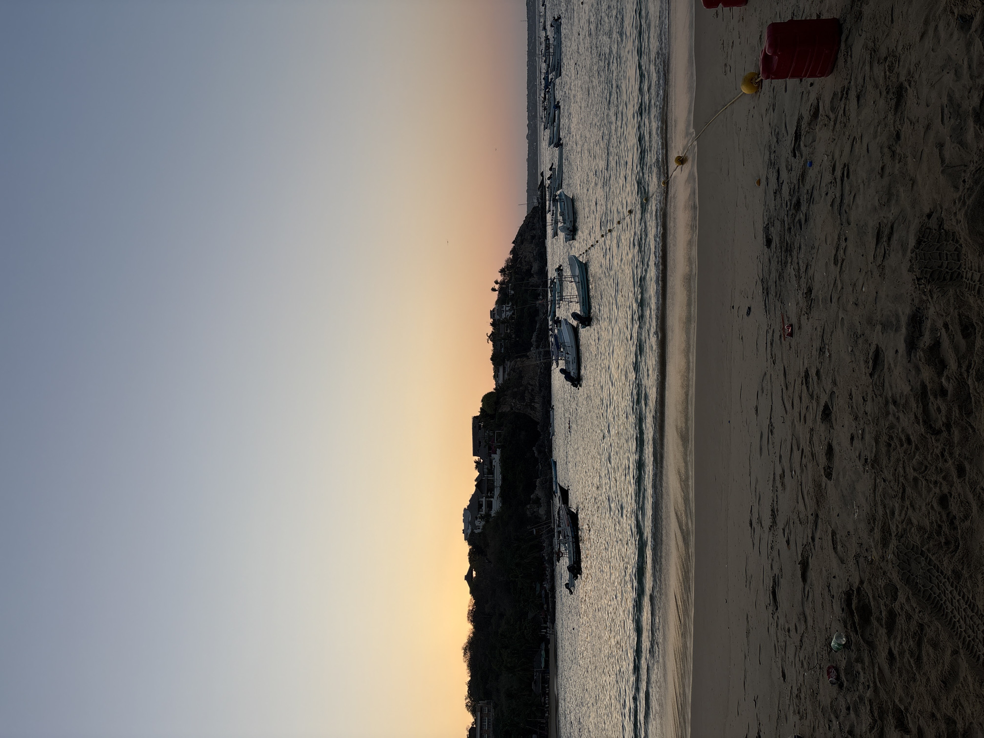The image depicts a coastal scene at sunset in Puerto Escondido, Mexico. The sky transitions from blue to orange, and the sun appears to be setting behind a coastal hill. Several boats are anchored in the water, which is separated from a dark sand beach by a narrow strip of light sand. A red container and yellow buoy are visible on the beach. Footprints are visible in the dark sand. The coastal hill is covered with trees and buildings.