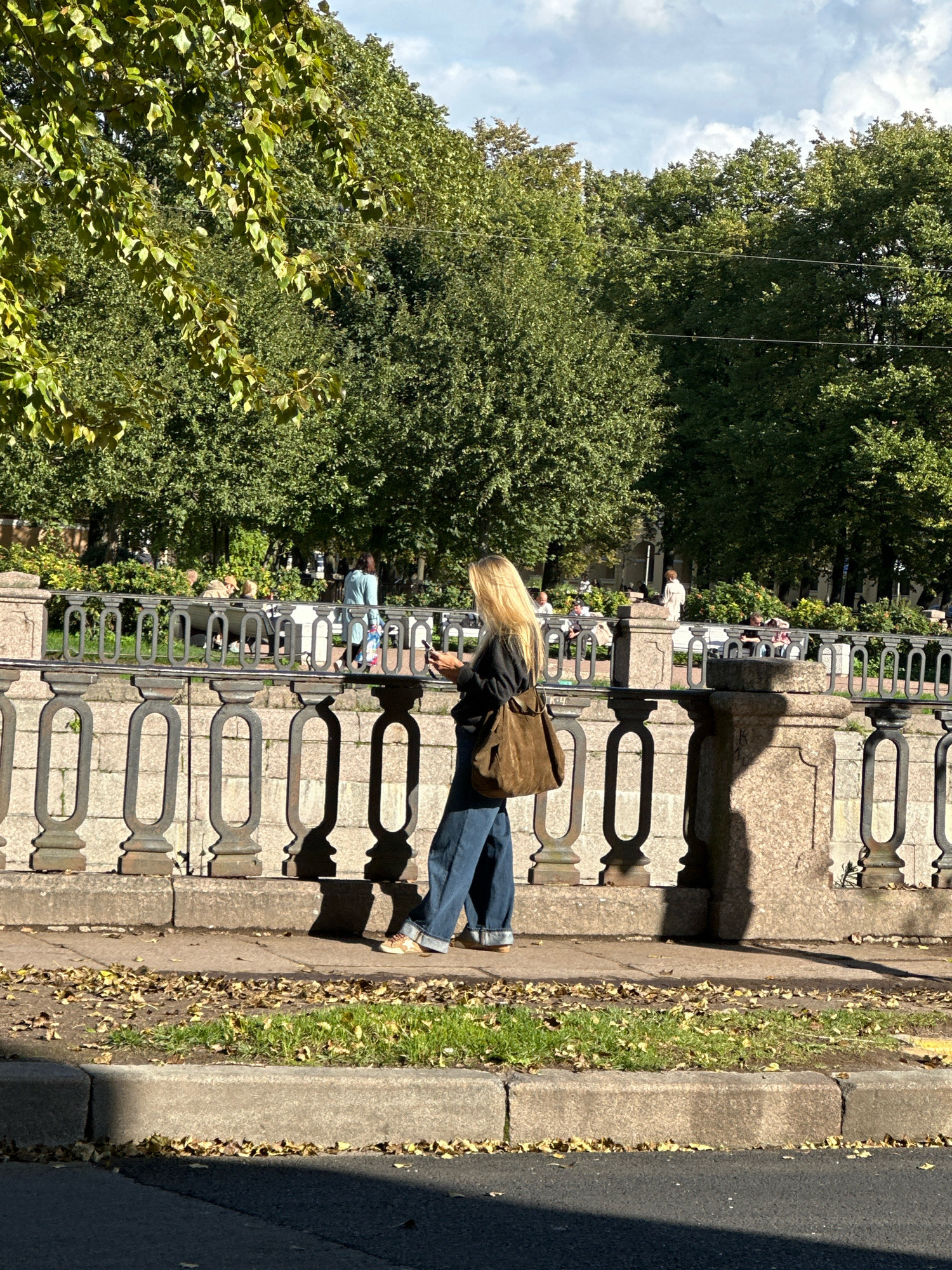 In this image, a blonde woman in blue jeans and a dark jacket walks away from the camera on a sidewalk, holding a mobile phone. She is carrying a brown canvas shoulder bag. The sidewalk is beside a stone embankment with a decorative metal railing. Beyond the railing, people are sitting on benches under trees in a park. The scene appears to be a sunny day with shadows cast on the embankment and sidewalk. The trees are full of green leaves, suggesting it is either late spring or summer. The architecture of the embankment and railing, along with the style of the park setting, are consistent with many canal embankments and public gardens in Saint Petersburg, Russia.