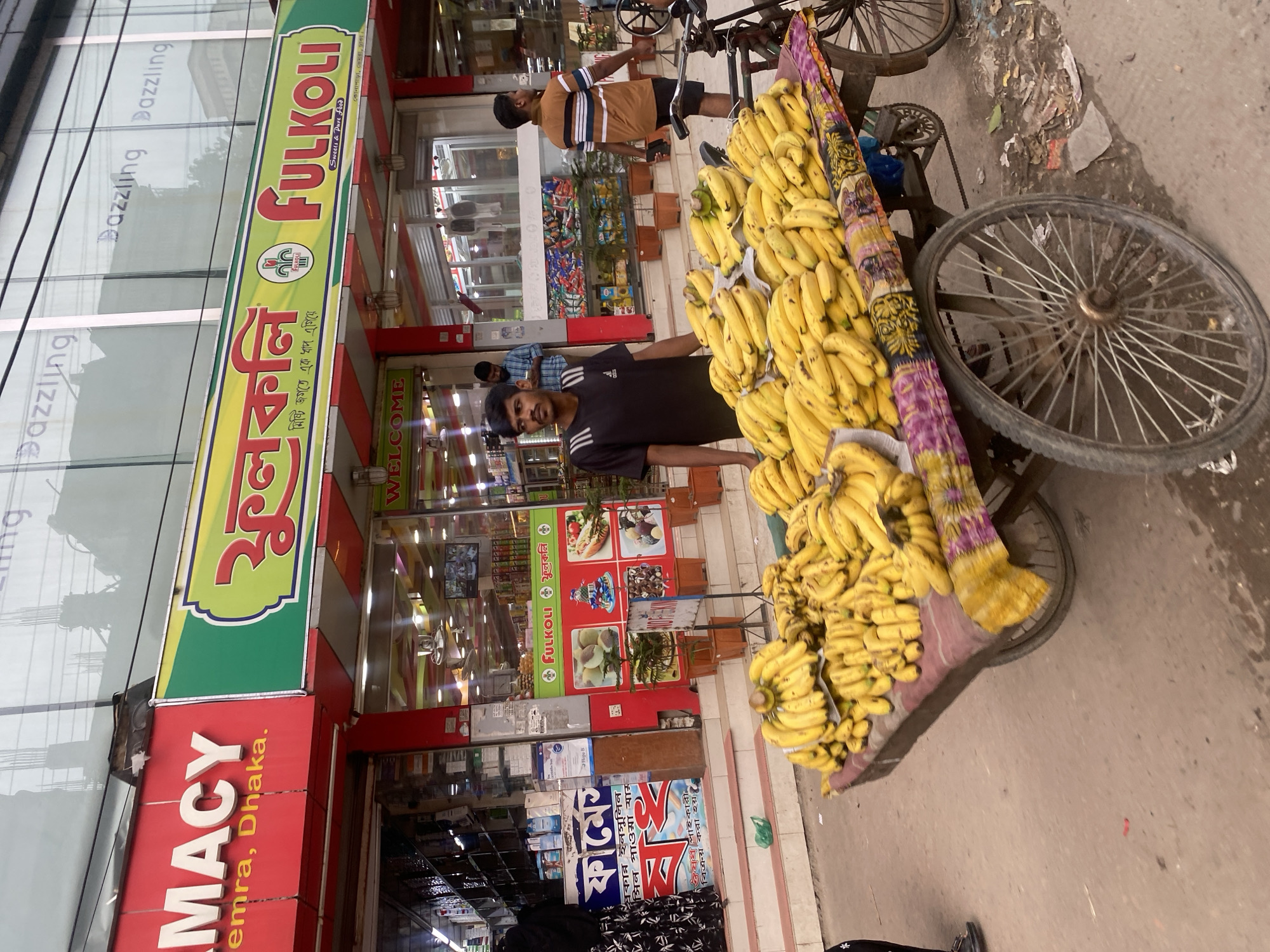 The image depicts a street scene in Dhaka, Bangladesh, likely in a commercial area. In the foreground, a large cart is overflowing with a significant quantity of ripe yellow bananas. The cart has two large spoked wheels and is decorated with a colorful, patterned cloth. Behind the cart, a young man with dark hair and a mustache, wearing a black t-shirt with white stripes on the sleeves, stands looking forward, appearing to be a vendor. To the right of the cart, a bicycle is parked.
In the background, there are storefronts. The building on the left has a red sign with white lettering that reads "EMACY" and below it "emra, Dhaka.". To the right of that, a vibrant green and yellow sign with Bengali text and the word "fulkolli" in English, along with "Sweets & Past Food" and smaller Bengali text, is prominently displayed. This sign suggests a sweet shop or confectionary. The storefronts display various goods and have glass entrances, revealing interiors with shelves stocked with products and ceiling fans. Another man, wearing a light brown t-shirt with horizontal stripes, is walking away from the camera in the background. The overall environment appears to be a busy urban street with storefronts and pedestrian activity.