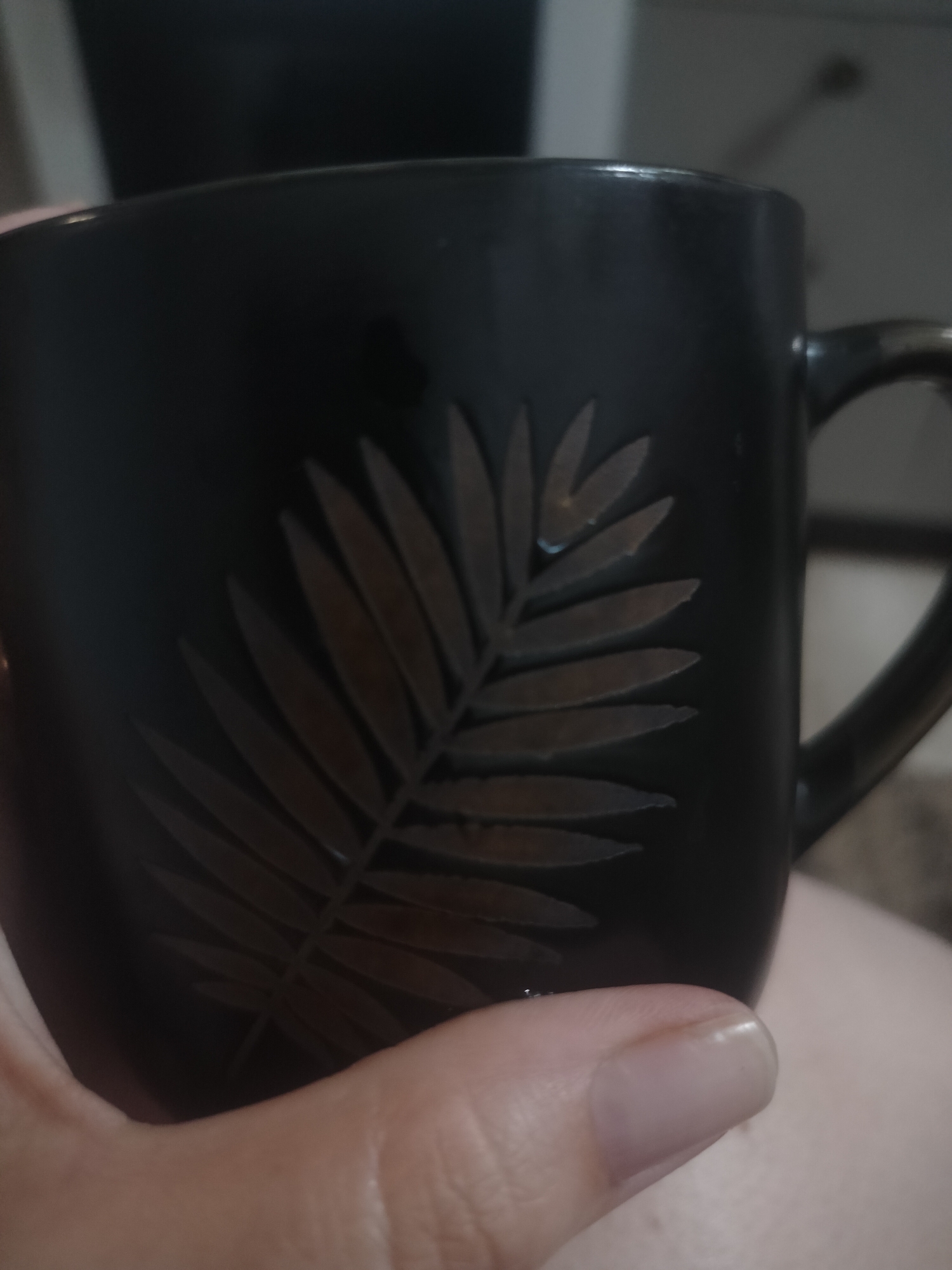 The image shows a close-up of a dark-colored mug with a raised leaf design on its surface. A hand is holding the mug, with a thumb and fingernail visible in the foreground. The mug is the primary object, and the hand is interacting with it. The background is dark and out of focus, providing no significant scene or setting details. There is no visible text in the image. The lighting appears to be indoor, as suggested by the soft illumination and lack of harsh shadows. The image does not convey any specific activity or event beyond the act of holding a mug. There are no indicators of time of day, weather, or specific location cues, beyond the provided "Yarovoye, Russia" context which is not visually evident. The overall mood is neutral.