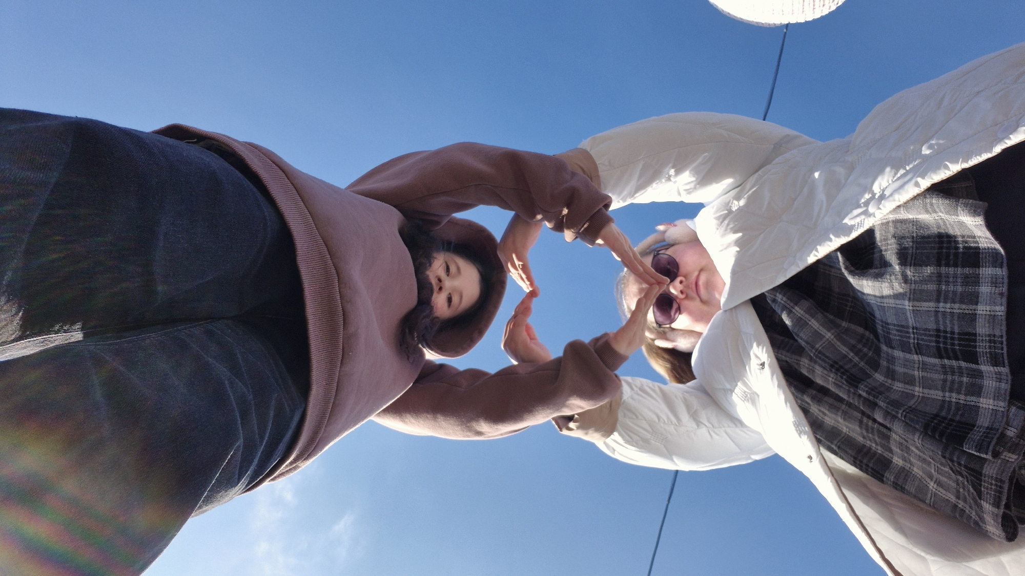 A low-angle, wide-shot photograph captures two young women forming a heart shape with their hands against a clear blue sky. The perspective looks up at the subjects, creating a slightly exaggerated sense of scale.
On the left, a woman with dark hair wears a brown hooded sweatshirt and dark pants, her face gently smiling as she looks down towards the camera. Her hands, meeting the other person's, contribute to the heart gesture.
On the right, another woman wears a white quilted jacket over a grey and black plaid shirt. She has sunglasses on her face, and her expression also suggests contentment. Her hands complete the heart shape in the center of the frame.
The background is an uninterrupted, bright blue sky, indicating a clear, sunny day. A faint, light-colored object, possibly a hanging decoration or lamp, is partially visible in the upper right corner of the frame. Rainbow lens flares are noticeable on the left woman's dark pants in the lower-left portion of the image. The scene takes place in Chaikovsky, Russia, conveying a moment of friendship or affection. No text is visible in the image.