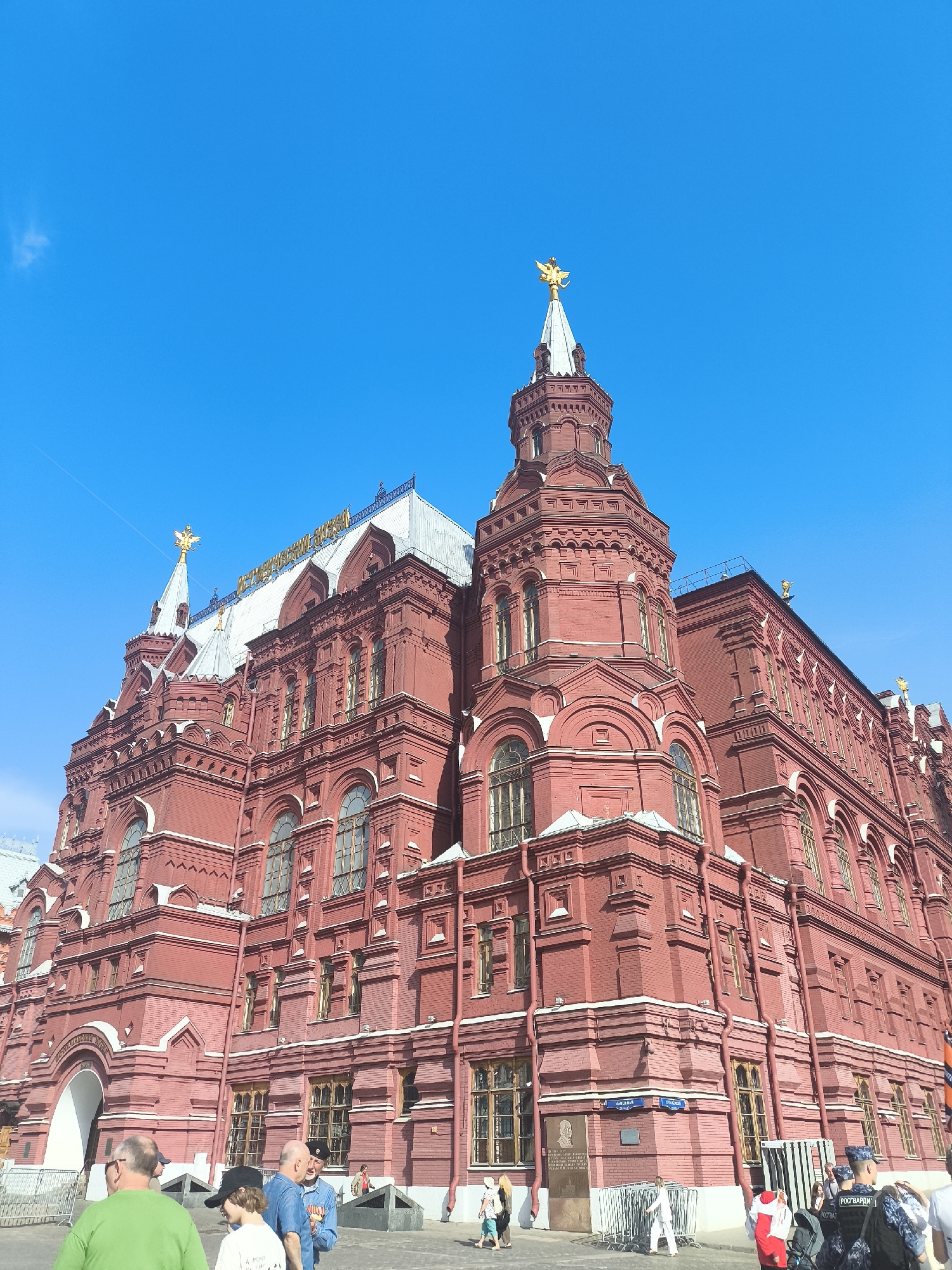The image shows the State Historical Museum in Moscow, Russia, viewed from a low angle on a sunny day. The museum is a large, red brick building with multiple levels, arched windows, and decorative architectural details. The roof has a silver-colored covering and is adorned with spires featuring golden eagle statues on top. A sign is visible on the roof. In the foreground, there are several people, some walking, some standing. A street sign is visible on the side of the building. The sky is a clear, bright blue.