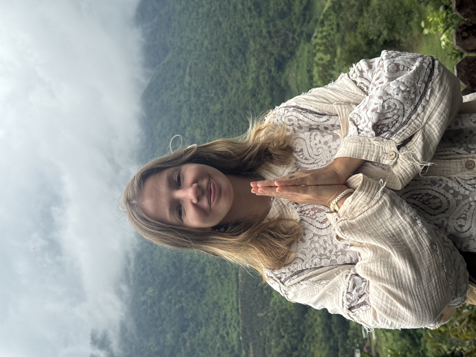 A medium shot captures a young woman with blonde hair and a light complexion from the chest up, standing outdoors with her hands pressed together in front of her chest in a prayer-like gesture. She is wearing a cream-colored, loose-fitting garment with lace accents. The background consists of lush, green, mist-covered mountains, suggesting a location in a mountainous or hilly region. The sky is overcast with grey clouds. The image appears to have been taken during the day. There are no identifiable landmarks indicating the city of La Dạ, Vietnam.