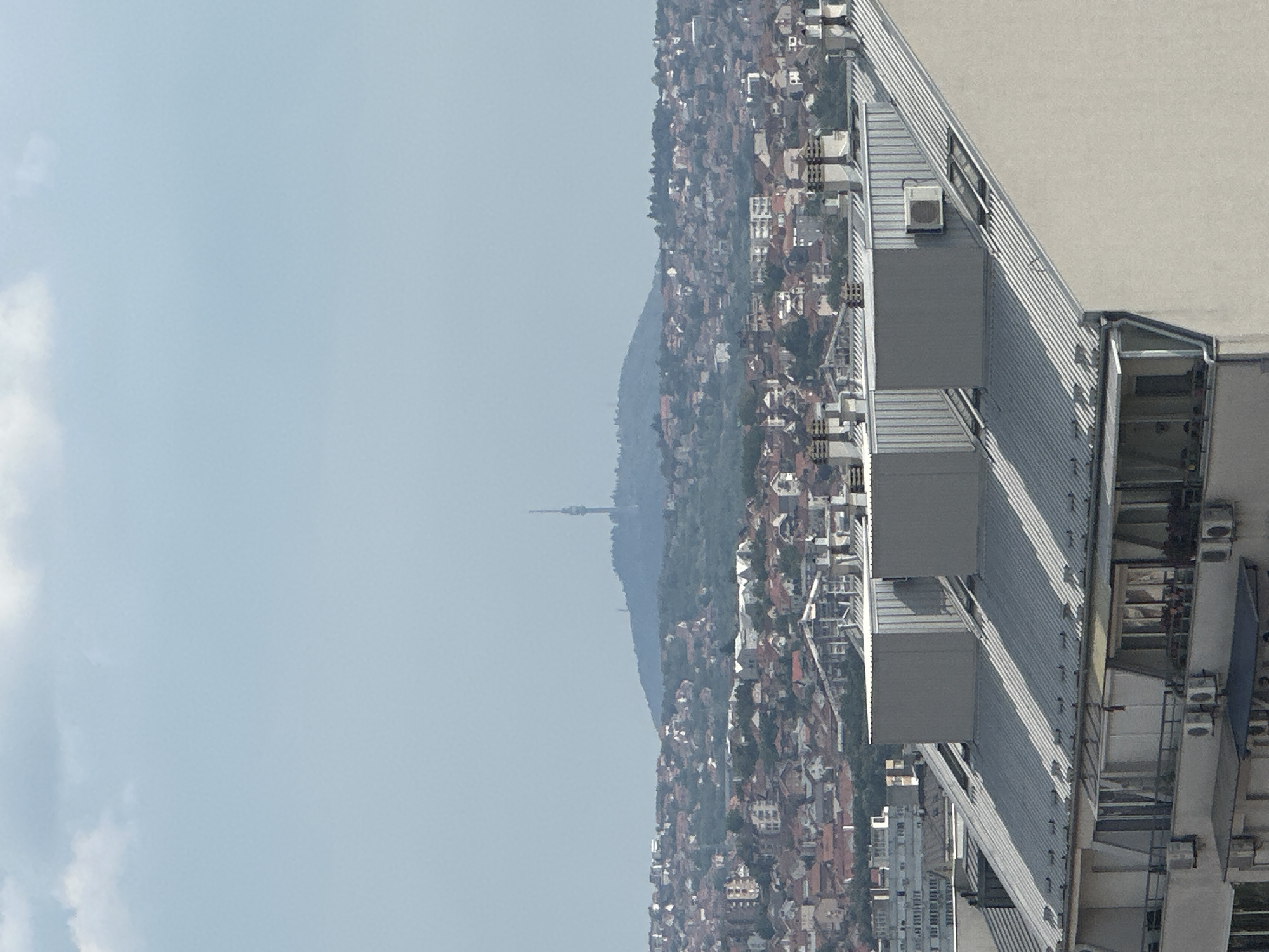 This is a vertically oriented, high-angle photograph of a cityscape, likely taken from a building in Belgrade, Serbia. The foreground is dominated by the roof and upper floors of a modern building, characterized by its metal siding, pitched roof sections, and visible air conditioning units. Balconies and windows are also apparent.
Beyond this structure, the image reveals a densely populated hillside covered in numerous buildings with terracotta roofs, indicative of a residential area. The buildings are clustered together, extending up the slope of a large hill. Atop the hill, silhouetted against a hazy sky, is a tall, slender tower, likely a television or communication tower, a prominent landmark in Belgrade.
The sky is a pale blue with some soft, white clouds visible on the left side of the frame. The overall lighting suggests it is daytime, possibly late morning or early afternoon, given the brightness and the angle of shadows cast on the building in the foreground. The atmosphere appears slightly hazy, which softens the details of the distant landscape.
There are no visible people or specific activities taking place that can be discerned from this distance and angle. The scene depicts a typical urban panorama, showcasing the juxtaposition of modern architecture with older residential areas on a hilly terrain. No text is visible in the image.
