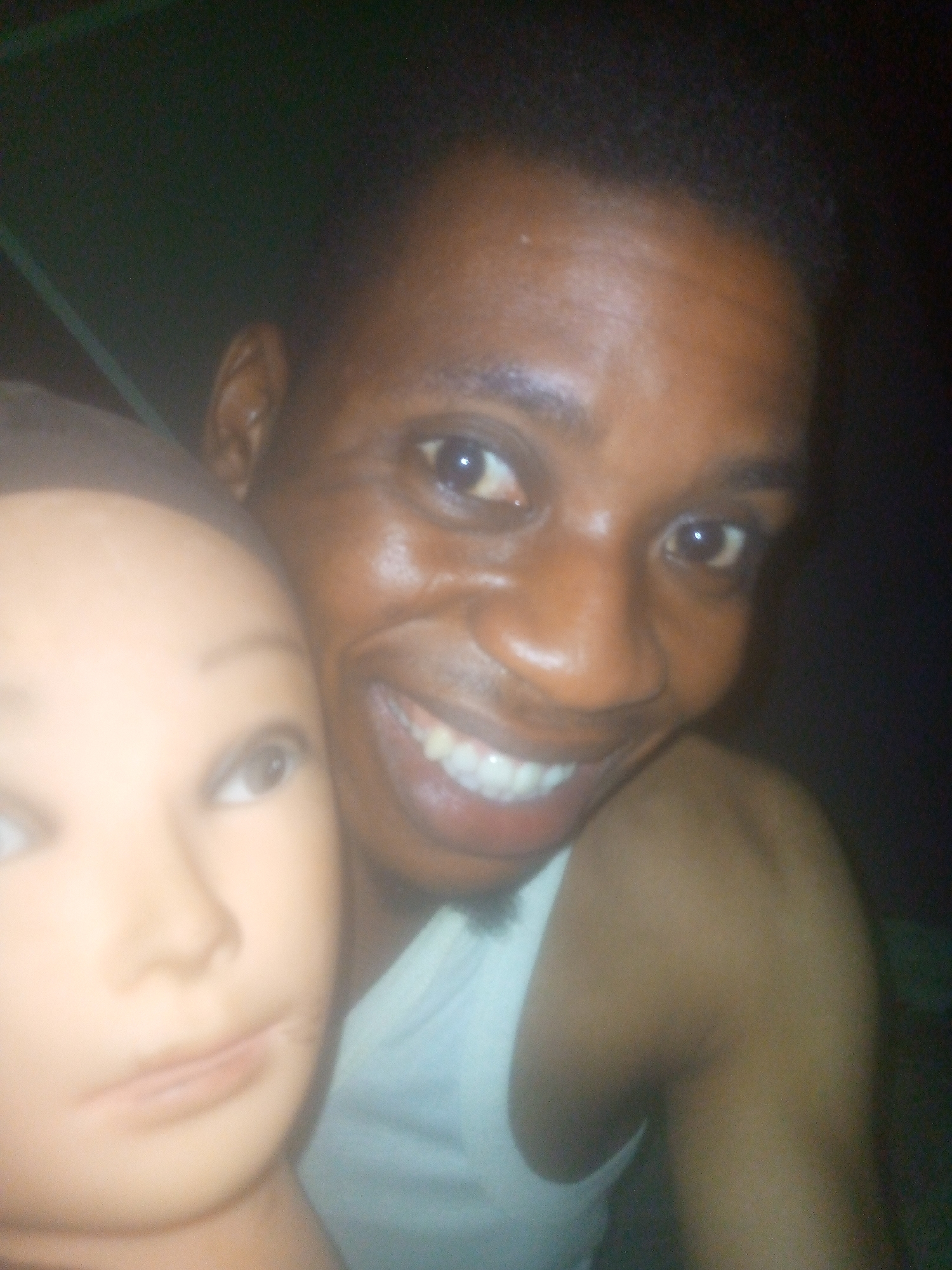 A man is holding a mannequin head. The man is smiling and wearing a white tank top. The background is dark. The location is unknown.