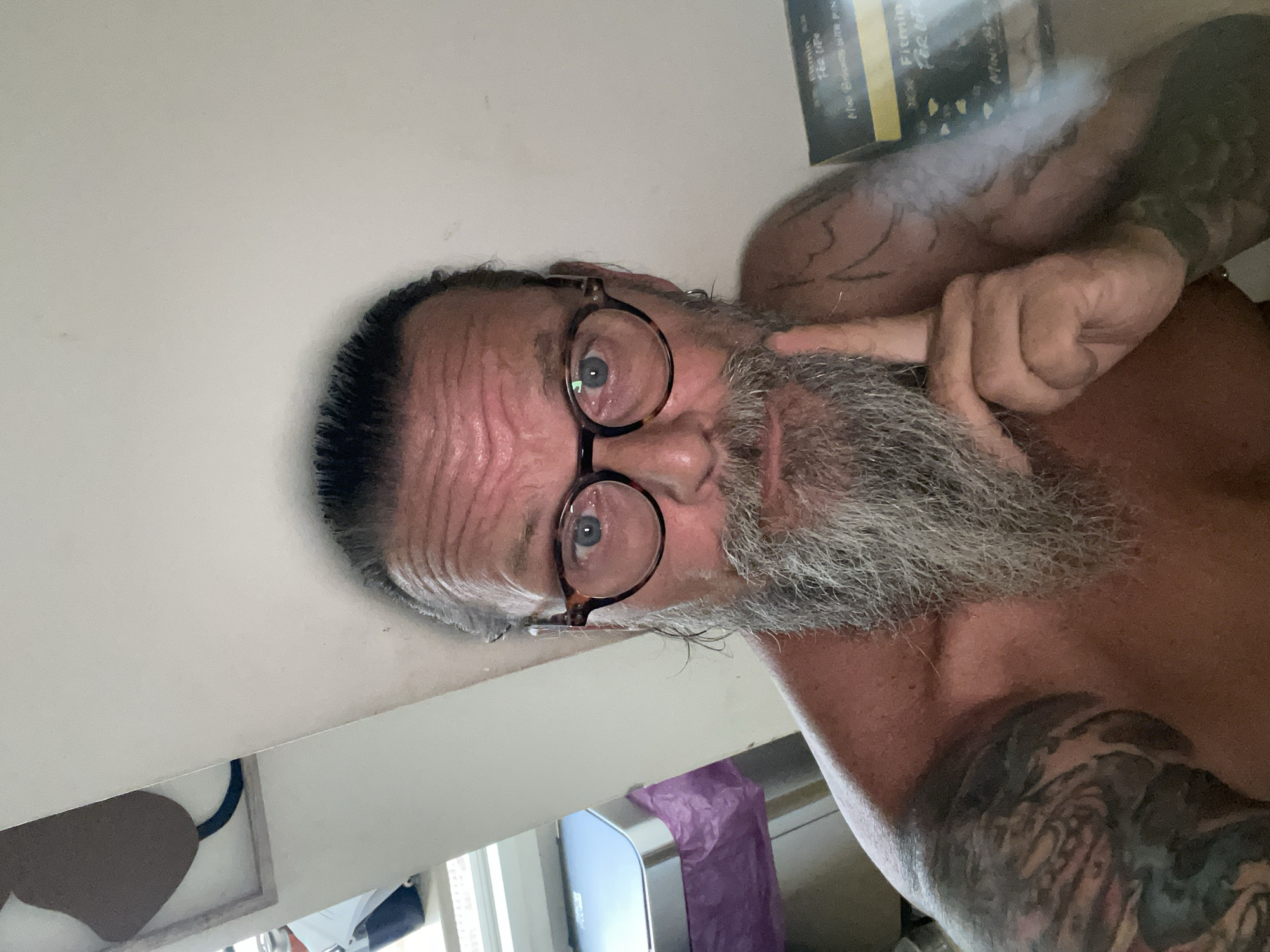 The media file is a close-up, portrait-style image of a man with a grey beard and tattoos. The man is wearing glasses with tortoiseshell frames and is looking directly at the camera. He has his right index finger extended, touching his cheek near his eye. His hair is dark and slicked back, with some strands falling onto his forehead, which appears to have wrinkles. His skin is fair, and his eyes are blue.
The man is shirtless, revealing prominent tattoos on his arms and chest. The background is blurred and consists of a white surface, possibly a wall or a desk, with some indistinct objects and shapes visible. One object that stands out is a yellow and black label or book spine in the upper right portion of the image, with some writing on it. The overall lighting suggests it could be daytime.
The image is oriented sideways, with the man's head towards the left of the frame. There are no clear indicators of the specific activity or event taking place, other than a posed portrait. The man's expression is neutral, possibly contemplative or amused. The visible text on the yellow and black object is too blurry to be read with certainty. The context provided states the location is Netanya, Israel.