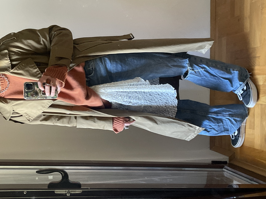 An individual is depicted standing indoors in Boreti, Montenegro, captured from mid-torso to their feet. The person wears a light tan trench coat over an orange knit sweater, wide-leg light blue distressed denim jeans with a rip on the left knee, and black Vans Old Skool sneakers with white soles. A white textured, possibly lace, fabric hangs from beneath the sweater over the jeans. The individual's right hand holds a smartphone encased in a camouflage-patterned cover; multiple rings, one with a reddish-brown stone, are visible on this hand. A silver-toned chain necklace is worn over the orange sweater. The person stands on a light-colored wooden plank floor. To the left, a dark-framed structure, resembling a door frame or mirror, with an attached dark hook, is present. The background consists of a plain, light-colored wall.