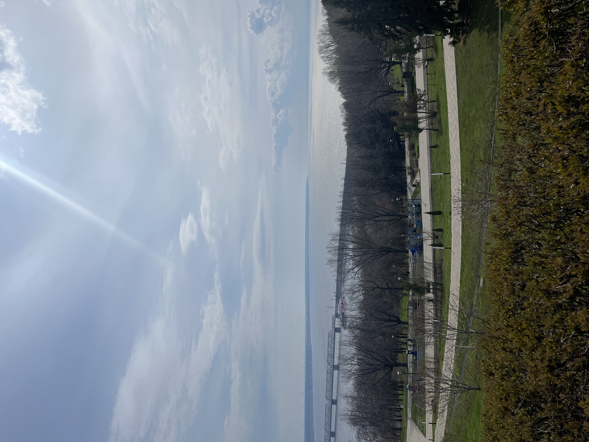The image captures a panoramic outdoor scene in Ulyanovsk, Russia, viewed in bright daylight. The primary features are a vast body of water, a tree-lined park, and a sky with scattered clouds.
In the upper portion of the frame, the sky is mostly clear and light blue, adorned with wispy white clouds. A prominent sun flare emanates from the top left corner, indicating the sun's position and contributing to the bright lighting conditions.
The middle ground is dominated by a wide expanse of water, likely the Volga River, which shimmers with reflected sunlight. In the distance, stretching across the river, a long, dark-colored truss bridge is visible, connecting the landmasses on either side. A faint haze hangs over the far bank of the river.
On the right side of the image, running parallel to the water, is a park or embankment area. This section features numerous deciduous trees, many of which are bare, suggesting early spring or late autumn. Several paved walking paths wind through areas of green grass. Along these paths, lampposts, park benches, and some blue playground equipment can be discerned. The immediate foreground on the far right is filled with a dense band of bushes or trees with green foliage, possibly evergreens or early-leafing varieties.
No people or specific activities are clearly visible in the image. The scene conveys a peaceful, open atmosphere under a bright sky. There is no visible text in the image.