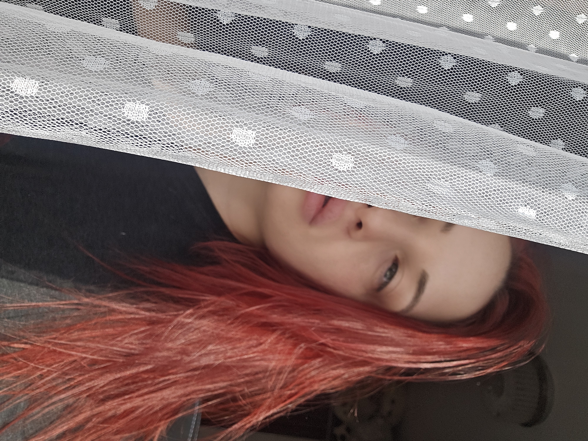 A person with dyed red hair lies partially concealed behind a white, polka-dotted mesh fabric. Only their face, from the bridge of their nose downwards, and their hair are visible. The person is wearing a dark-colored top. The background is dark and blurry. The image is rotated. The location is not discernible based on the given image.
