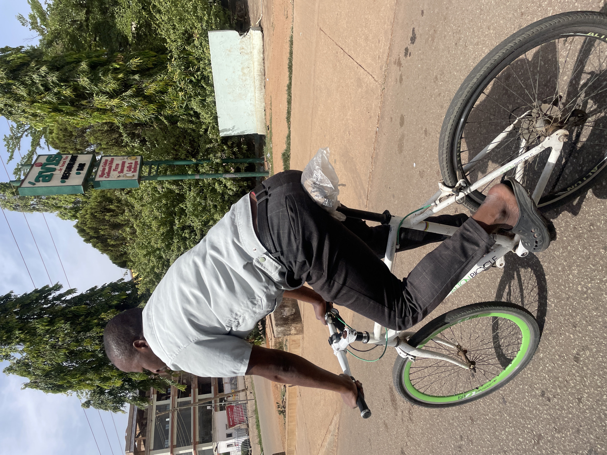 A man rides a bicycle on a paved road in Jos, Nigeria. He is wearing a light grey, short-sleeved shirt, dark pants, and black slip-on shoes. He is carrying a plastic bag. The bicycle is white with a green front tire rim. A sign for "Ams Restaurant & Bar" is visible in the background, along with trees and buildings.