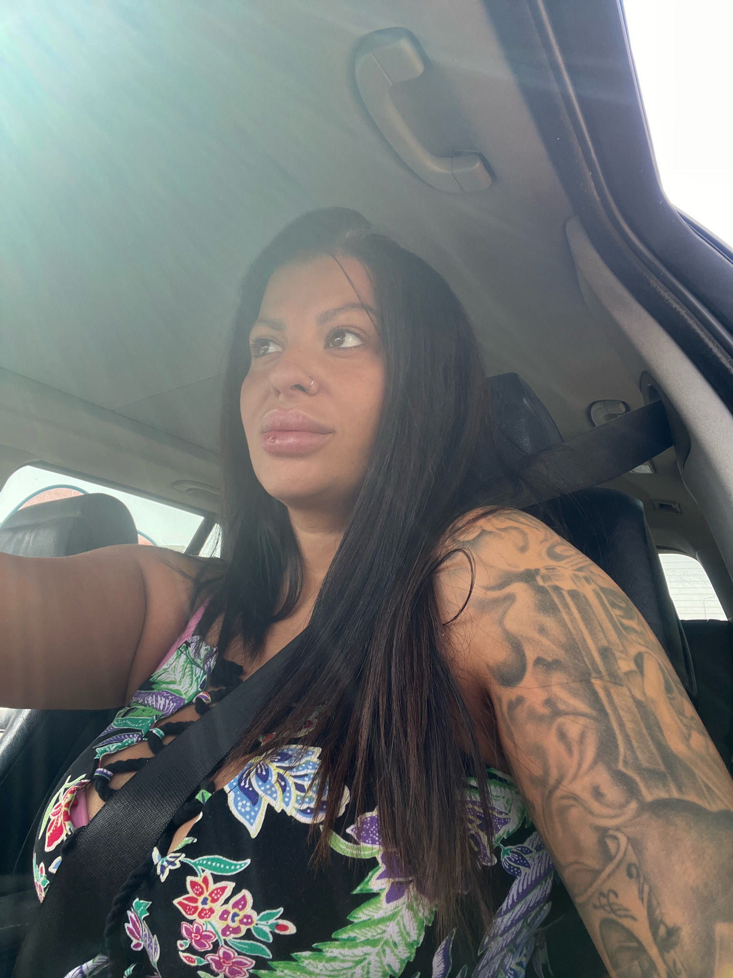 An adult woman with long dark hair and a nose ring is pictured from the shoulders up, sitting in the driver's seat of a car. She is wearing a black floral print top with cutouts and a seatbelt across her chest. Her left arm, visible in the foreground, is covered in a large tattoo. The interior of the car is visible, including the headliner and a grab handle. The scene is outdoors, as suggested by the sunlight streaming into the car. The image does not contain enough information to determine the specific location within the city of Plovdiv, Bulgaria.