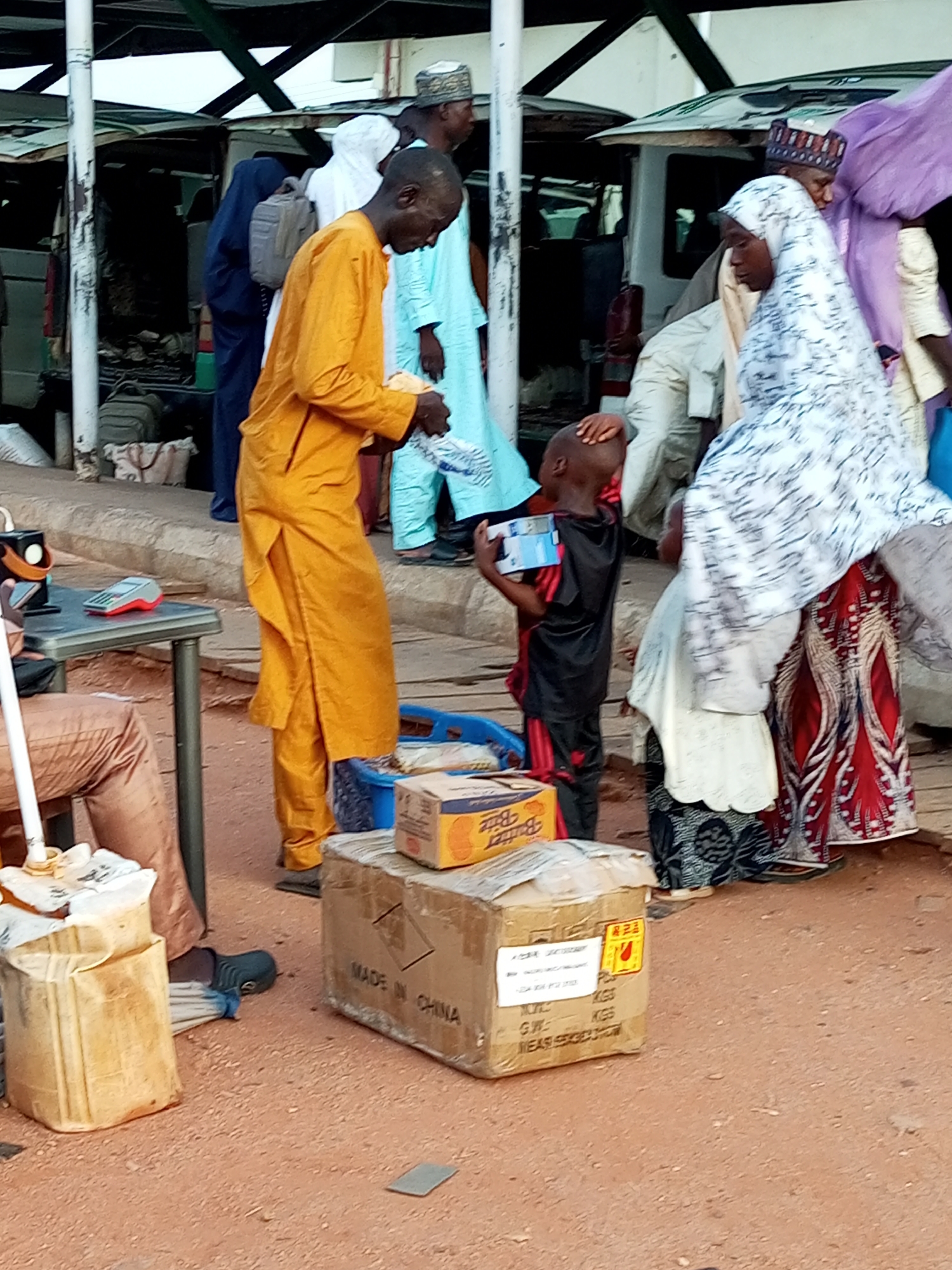 The image depicts a bustling outdoor scene in Gombe, Nigeria, likely at a marketplace or transport hub. In the foreground, a child in a black and red outfit is holding a small box, looking upwards. To the left, a man in a bright orange traditional garment stands near a small table with a calculator and a white jug-like object. In the background, several individuals are visible, some wearing traditional attire and head coverings, and various vehicles, including vans, suggest ongoing activity and possibly travel. The ground is covered in reddish-brown dirt, with some cardboard boxes and other items scattered around, indicative of commerce and movement. The overall atmosphere appears lively and possibly crowded.