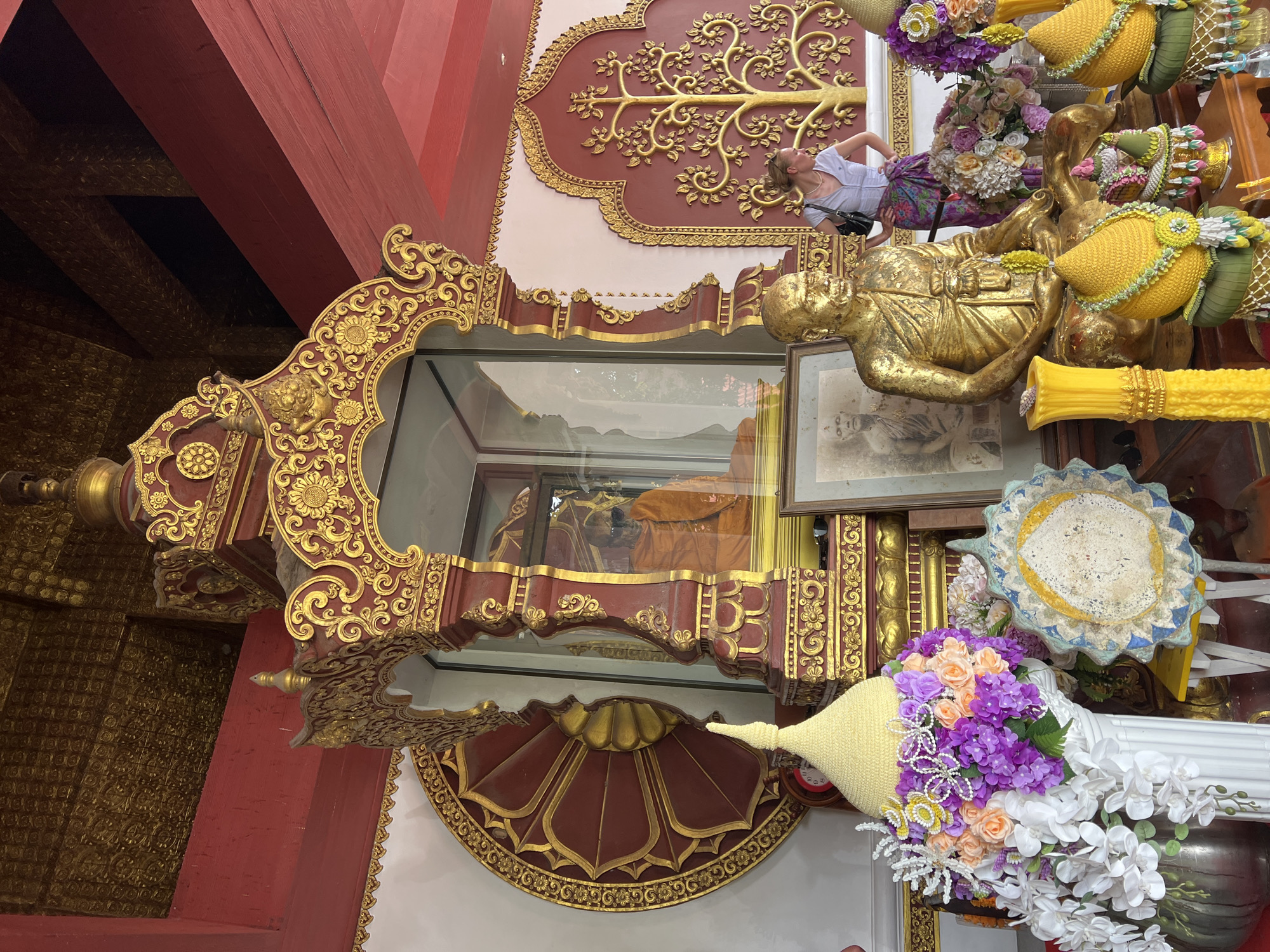 The image depicts an ornate shrine in Baan Hua Thanon, Thailand, likely a Buddhist temple or a place of religious significance. The focal point of the shrine is a glass-encased figure, possibly a revered monk or a statue of Buddha, draped in saffron robes. This sacred object is housed within an elaborate, gilded wooden structure adorned with intricate carvings and decorative motifs.
Surrounding the central display are various religious offerings and decorative elements. A golden statue of a seated figure, possibly another representation of a revered individual, is placed to the right, adorned with yellow, cone-shaped items wrapped in green and white fabric, suggestive of offerings. Several bouquets of artificial flowers, in shades of purple, pink, and white, are arranged around the shrine, adding a splash of color to the scene.
In the background, a young woman is visible, looking upwards at a decorative mural depicting a golden tree of life on a reddish-brown wall. Her posture and gaze suggest curiosity or reverence. The architectural style, with its deep red painted beams and intricate gold detailing, points to a traditional Southeast Asian aesthetic. The overall ambiance is one of solemnity and devotion, conveyed through the religious imagery and the act of offering. The lighting appears to be natural, suggesting it is daytime, but the weather cannot be determined from the image. No visible text is present in the photograph.