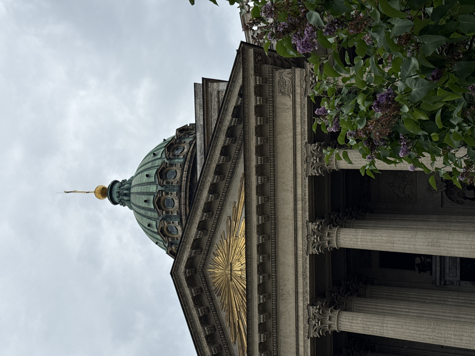 A low-angle, full shot captures the Kazan Cathedral in Saint Petersburg, Russia, under a cloudy sky. The green copper dome, topped with a gold orb and spire, is prominent in the upper portion of the frame. Below the dome, the classical facade of the cathedral is visible, featuring Corinthian columns and ornate architectural details, including a golden sunburst motif on the pediment. In the foreground, a cluster of blooming lilac bushes with purple flowers partially obscures the lower part of the cathedral. The scene is devoid of any people, indicating the time of day is likely not during peak tourist hours or the image was captured with no one in frame. The overall atmosphere is somewhat somber due to the overcast sky and muted colors.