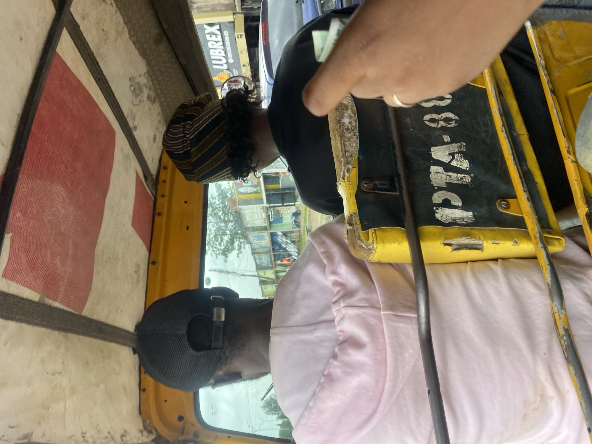A three-quarters shot from inside a yellow auto-rickshaw in New Stadium, Nigeria, shows two individuals from behind. The driver, wearing a black baseball cap and a light pink hoodie, is in the foreground. To their right, another passenger wears a striped cap and a black t-shirt. The auto-rickshaw's interior is visible, with a yellow ceiling adorned with red and white stripes. A portion of the vehicle's exterior, including the registration plate "PTA-88", is also in view. Through the windows, a street scene unfolds with buildings and a blue car. The image captures a mundane moment of travel within the city.