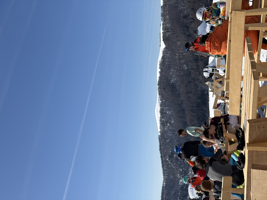 The image displays an outdoor scene at a mountain recreation area in Vranjak, Montenegro. A bright blue sky with a faint, linear contrail dominates the upper portion. The background features extensive dark, bare deciduous forests covering mountain slopes, topped by white, snow-covered peaks. The mid-ground reveals several small, dark-roofed wooden structures, partially buried in snow. The foreground is occupied by multiple light-colored wooden picnic tables with attached benches, set upon a snow-covered ground.
Approximately 10-15 individuals are present. Several are seated at the tables; these include a person in a dark grey and black athletic top, a child in a brown beanie, a person with dark hair and sunglasses on their head leaning forward, an individual in an orange jacket with their back to the viewer, and another in a colorful striped jacket and white helmet looking at a mobile device. Objects such as gloves and white mugs are visible on the tables. Other individuals are standing in small groups; one wears a blue helmet and dark jacket, another a light green t-shirt, and a third a red jacket and green goggles. A person in a red ski jacket and black helmet is seen holding skis. The scene indicates a social gathering, with people resting, conversing, and using electronic devices in a snowy mountain environment.