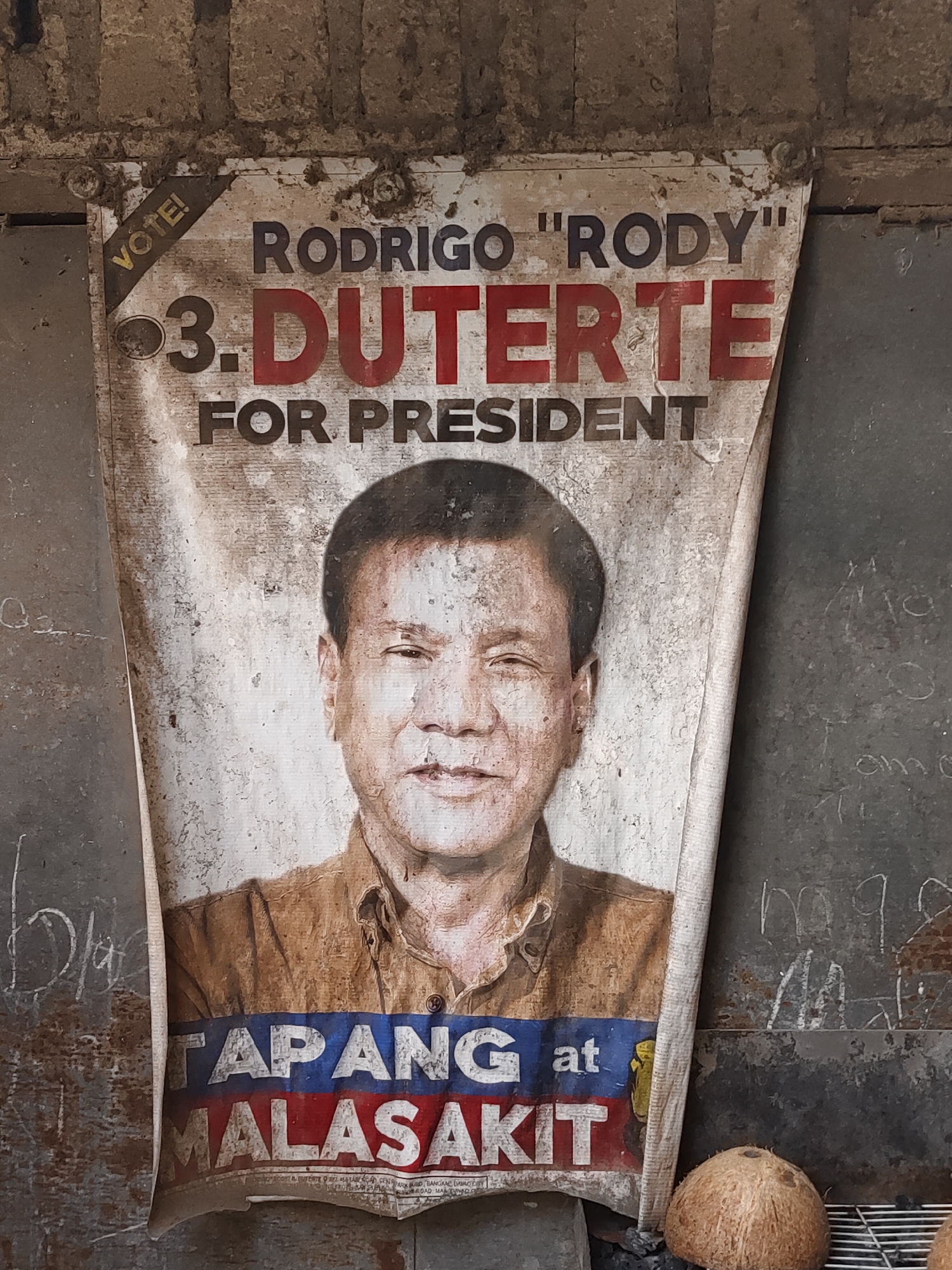 A weathered campaign poster for Rodrigo "Rody" Duterte for President is prominently displayed on a textured, dirty wall. The poster, which is torn and creased, features a portrait of Duterte. The text on the poster reads "RODRIGO 'RODY' 3. DUTERTE FOR PRESIDENT" and below his image, "TAPANG at MALASAKIT". The background wall appears to be made of rough, possibly concrete or brick, material with smudges and what looks like chalk or paint markings. To the right of the poster, there are coconuts and part of a metal grate visible. The overall environment suggests an outdoor setting in a rustic or unkempt urban area, consistent with locations in Davao City, Philippines, where Duterte served as mayor.