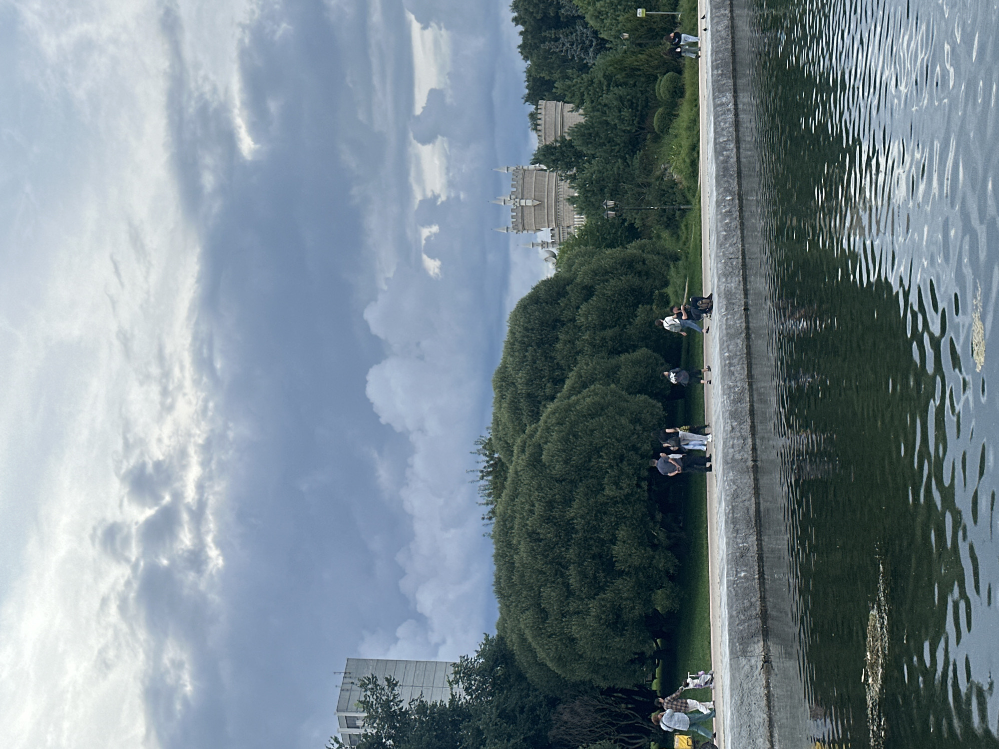 The image depicts a park scene in Moscow, Russia, likely Tsaritsyno Park. In the foreground, a body of dark water with ripples occupies the lower half of the frame. A concrete embankment runs along the edge of the water, separating it from a pathway. Several people are walking and standing on the pathway. To the left, a tall, light-colored building with a modern architectural style is visible above a cluster of trees. In the middle ground, large, rounded green trees dominate the landscape. Further in the background, a light-colored, castle-like structure with turrets and crenellations is partially obscured by dense green foliage. The sky is filled with dramatic, dark grey clouds, suggesting overcast weather. The overall atmosphere is serene, with a hint of grandeur from the distant architecture.
