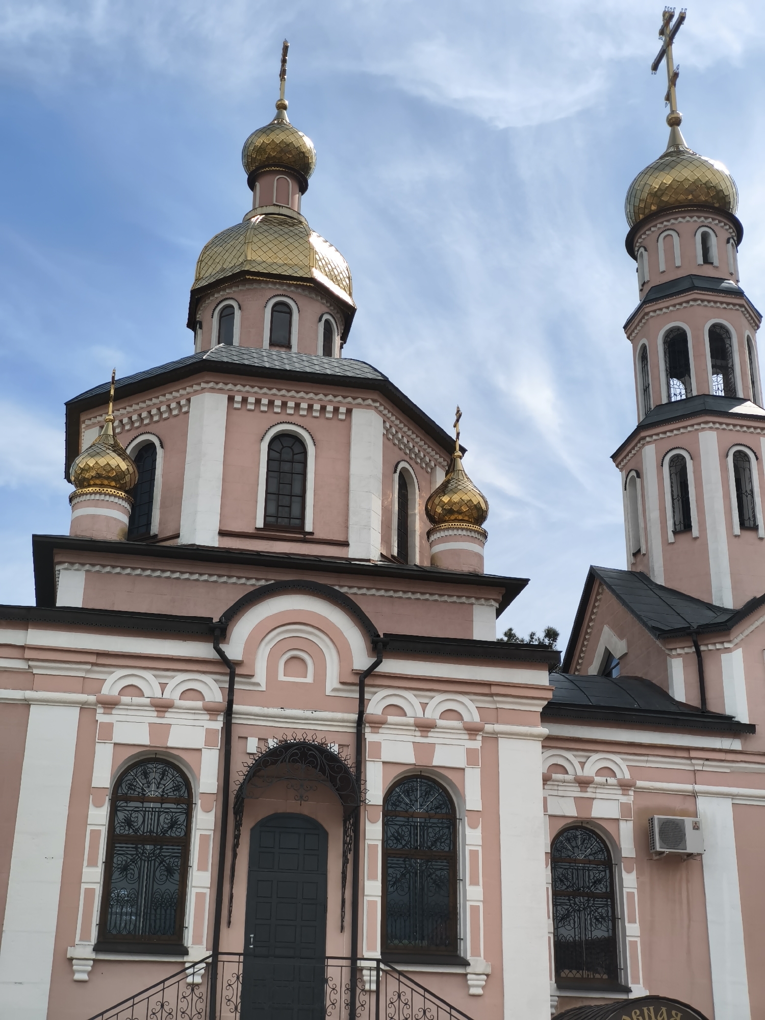 The image shows an exterior view of an Orthodox church located in the city of Arkhipo-Osipovka, Russia. The church is painted in pink and white with gold domes. It features a main building with a large central dome and several smaller domes, as well as a tall bell tower. The architecture includes arched windows and decorative elements. An air conditioning unit is visible on one side of the building. The sky is partly cloudy.
