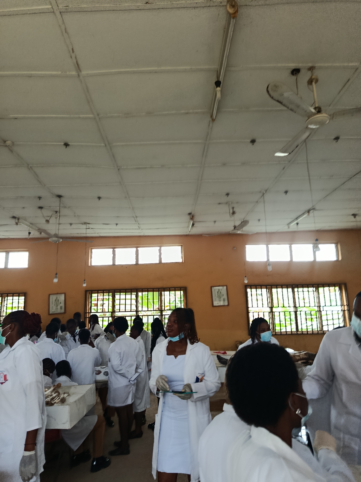 In an indoor classroom setting, a group of individuals, predominantly young adults wearing white lab coats and some wearing face masks, are gathered around tables. The room is sparsely furnished with a tiled ceiling, fluorescent lighting, and windows with bars. A ceiling fan is visible, suggesting a warm environment. The individuals appear to be engaged in a practical learning session, possibly in a science or medical field, as evidenced by the lab coats and the presence of what appears to be anatomical models or specimens on the tables. The overall atmosphere is one of focused activity and instruction.