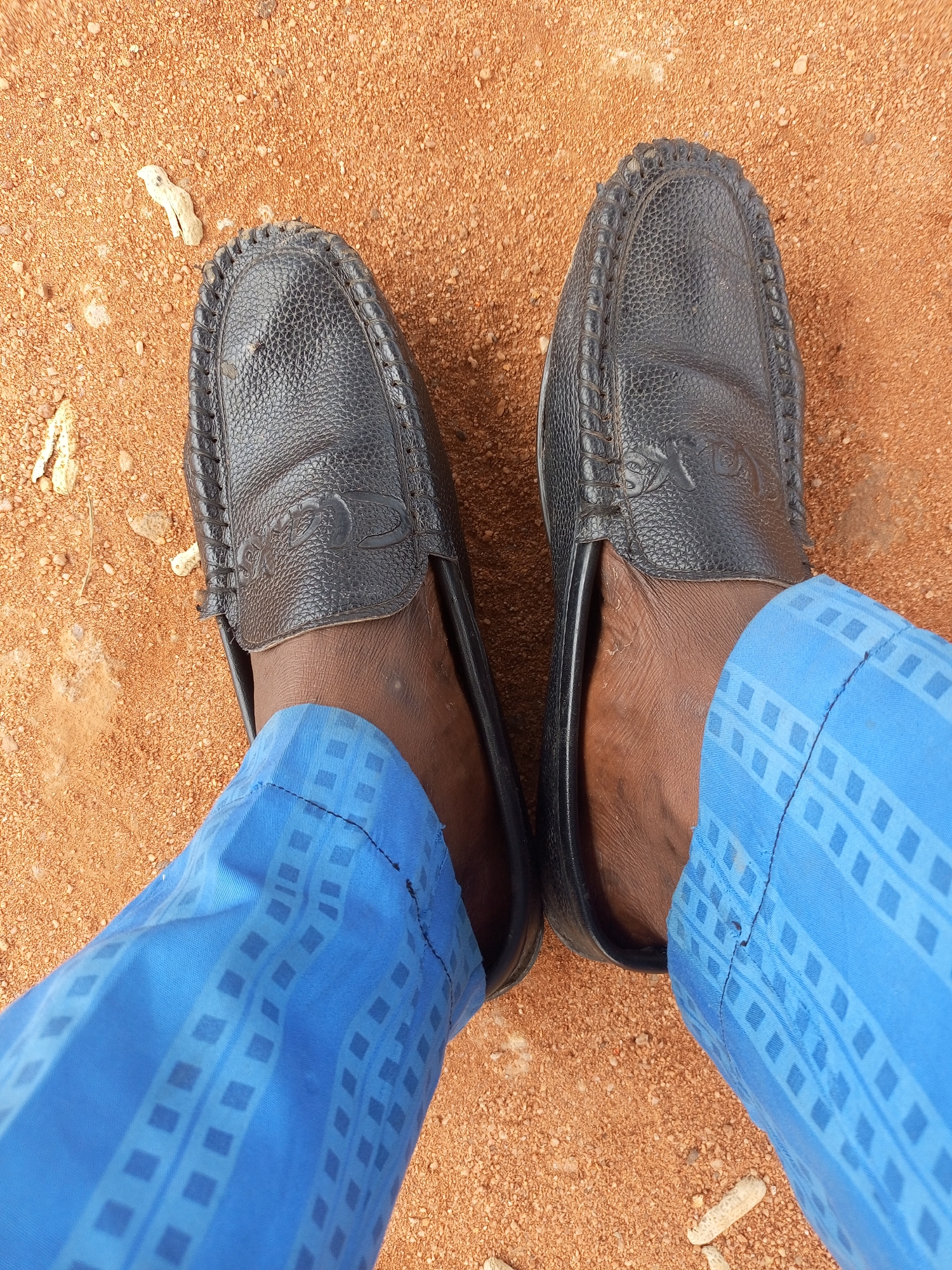 The image shows a close-up, overhead view of a person's feet, clad in black leather moccasins and bright blue patterned trousers. The shoes are resting on a surface of reddish-brown dirt and small stones, typical of unpaved ground, possibly a street or pathway in Tempure, Nigeria. The trousers feature a repeating geometric pattern of light blue squares within darker blue rectangles, creating a visually striking contrast with the earthy surroundings and dark footwear. There are a few small, pale organic debris pieces scattered on the dirt. The overall impression is of a casual outdoor setting.