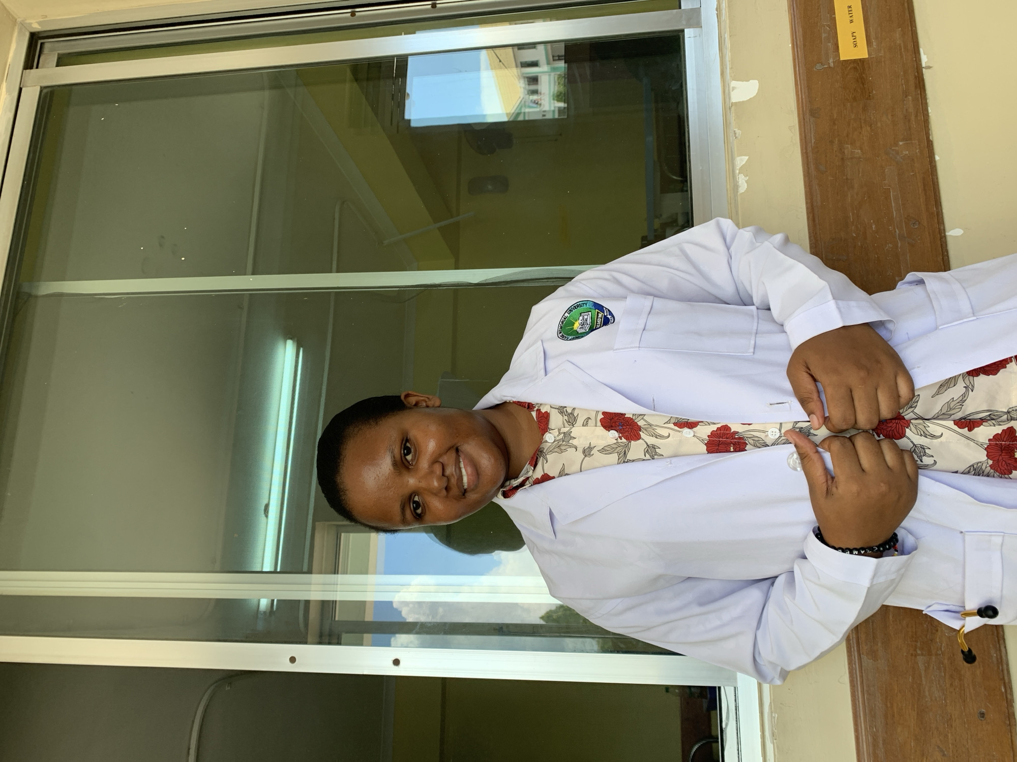 A woman in a white lab coat smiles at the camera. She is standing near a window with a reflection of a building and the sky. She wears a floral patterned shirt underneath the lab coat. Her hands are clasped in front of her, with a bracelet visible on one wrist. A brown wooden board with the words "SOAPY WATER" is partially visible on the right side of the frame. The location is potentially in Dar es-Salaam, Tanzania, based on the patch visible on the lab coat, but this cannot be verified with certainty from the image alone.