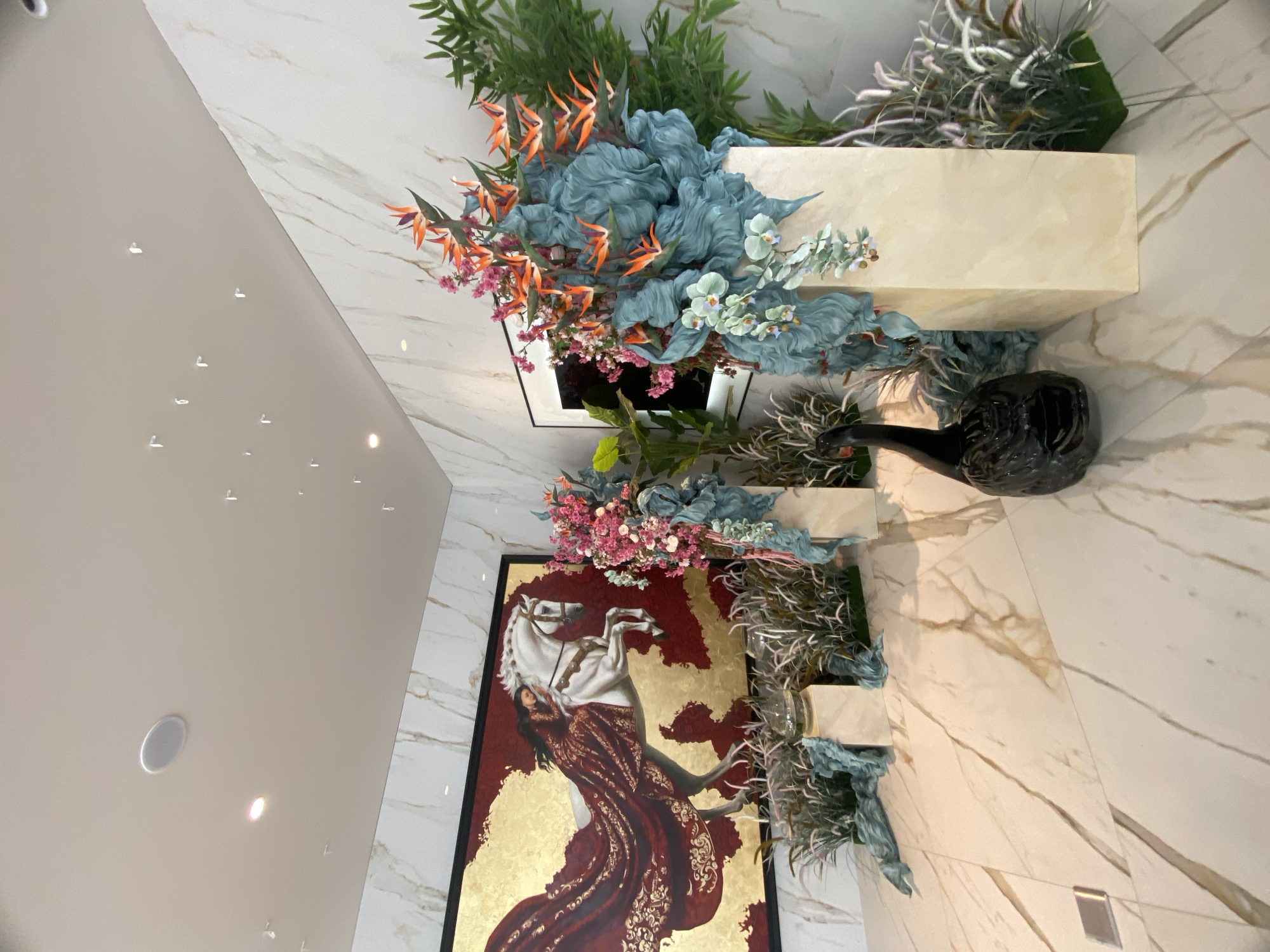 The image shows an indoor space with a marble-like wall and floor. A painting of a woman on a white horse dominates the scene, framed in black and featuring a mix of gold and burgundy colors. Above and around the painting and what appear to be display platforms are floral arrangements with green foliage, blue and orange flowers, and other decorative elements. There is a black object in the center. The space is well-lit, and a portion of the ceiling is visible with recessed lighting fixtures. There is no evidence to identify this space as located in Sochi, Russia.