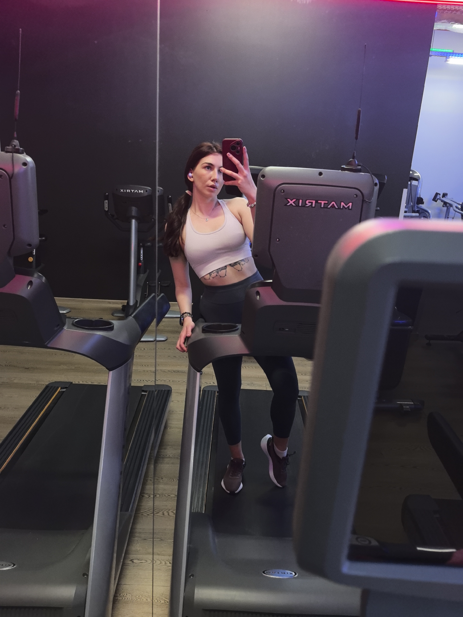 In a gym in Stara Zagora, Bulgaria, a young woman takes a selfie using her phone, while standing on a Matrix treadmill. She is wearing a light pink sports bra, dark leggings, and sneakers. She has brown hair, pulled back in a low ponytail, and is wearing a necklace and a bracelet. A second Matrix treadmill is visible to her left. A black wall is in the background, with a neon light above it. Other gym equipment can be seen in the background.