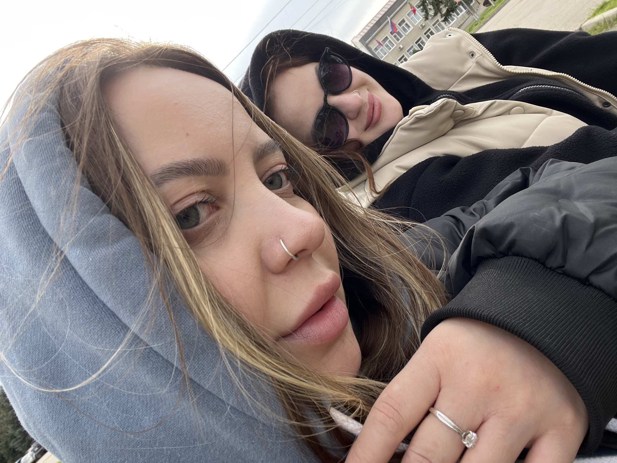 Two young women are posing for a selfie. The woman in the foreground has light brown hair and is wearing a gray hoodie. She has a nose ring and a ring on her left ring finger. The other woman is wearing a black hoodie, sunglasses, and a cream and black jacket. In the background, there is a building with the Russian flag and other flags. This image was taken in the city of Novopokrovskaya, Russia.